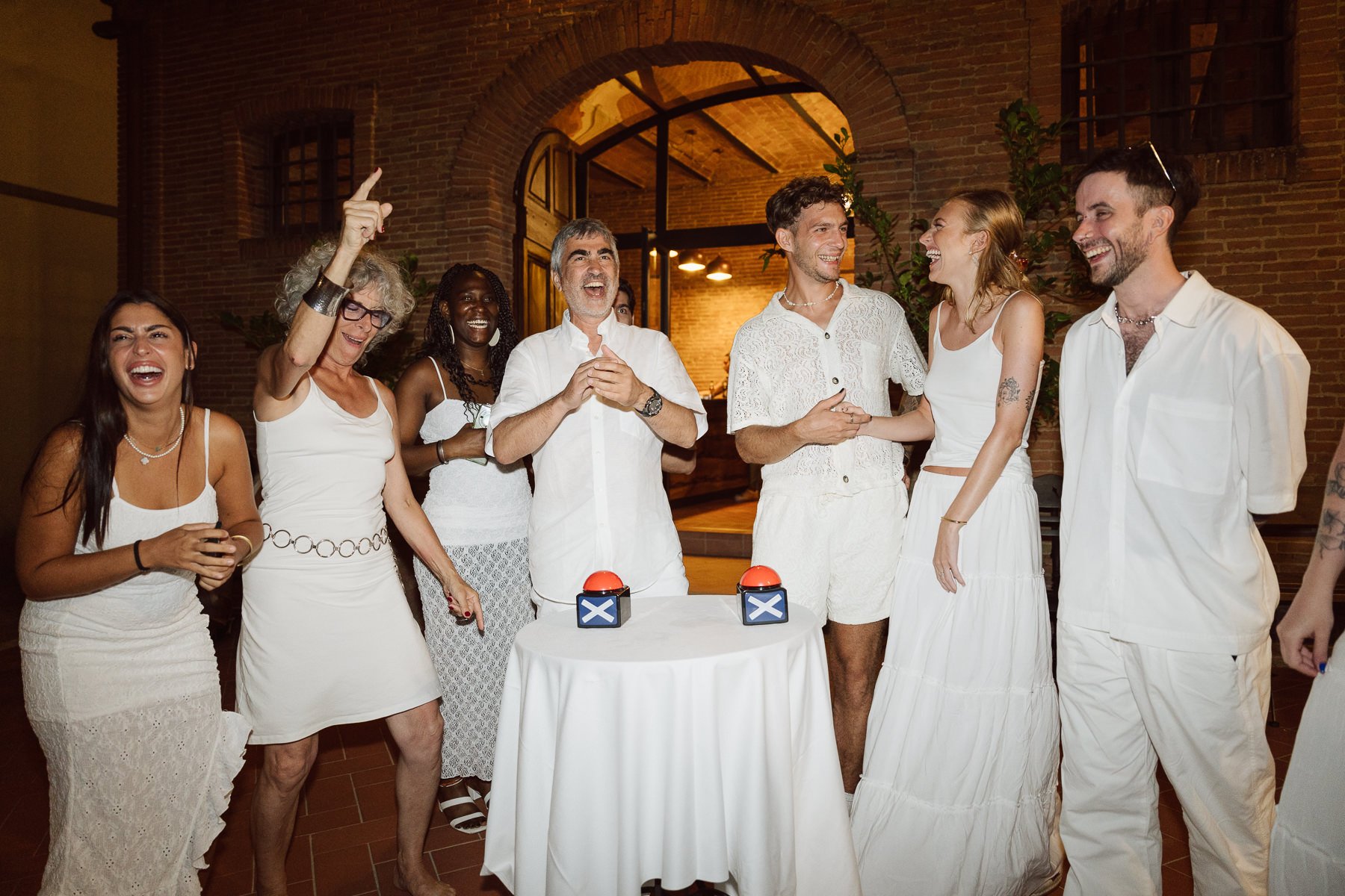 Invités en tenue blanche riant autour de buzzers rouges lors d’un quiz musical pendant la soirée de mariage en Italie.