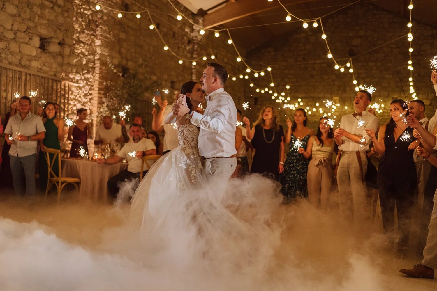 Les mariés ouvrent le bal lors de leur réception de mariage, entourés d’invités tenant des fontaines lumineuses, avec effet de fumée au sol et guirlandes lumineuses en arrière-plan.