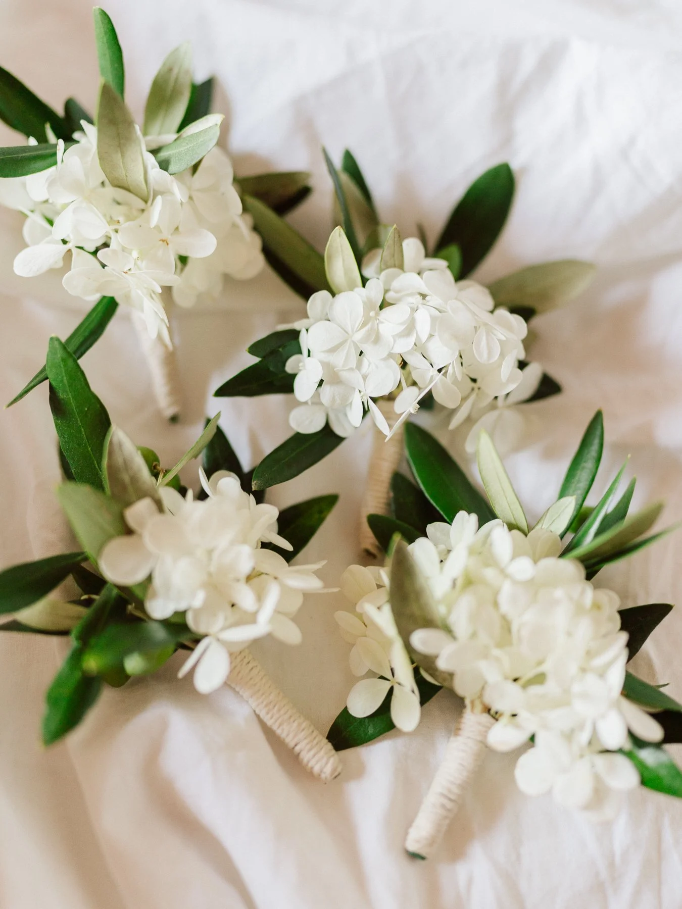 Boutonnières de mariage en fleurs blanches et feuillage d’olivier posées sur un tissu clair, détail élégant d’un mariage en Toscane