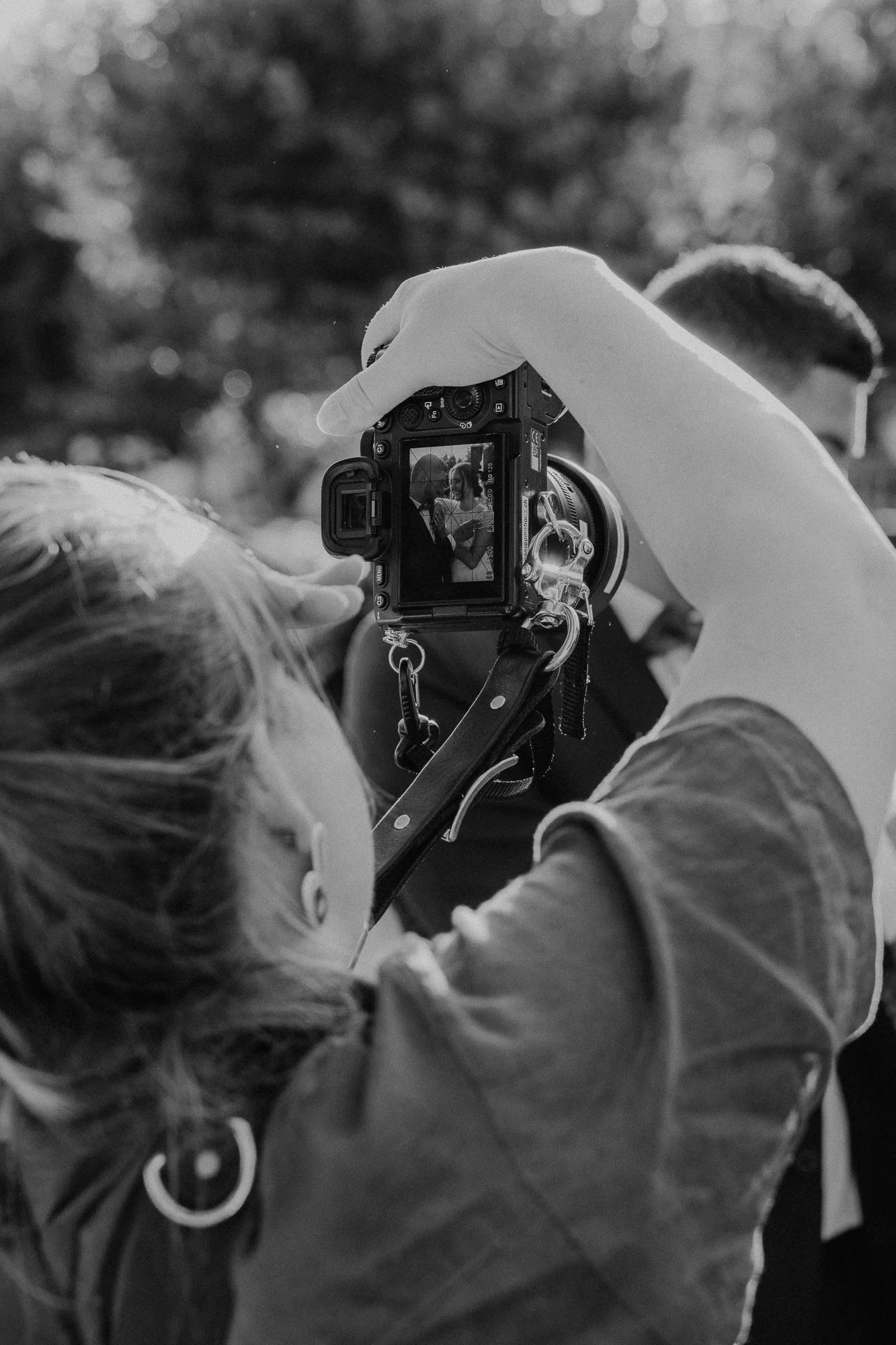 Une personne prenant une photo avec un appareil photo numérique, vue de dos, avec d'autres personnes en arrière-plan, en plein air, en noir et blanc.