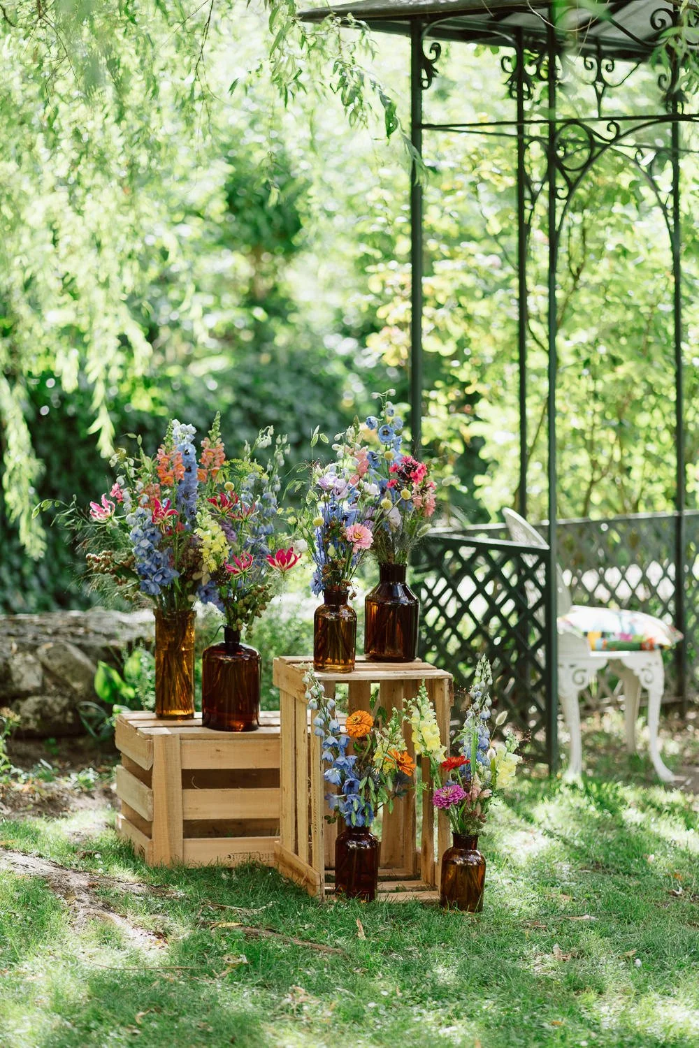 Fleurs colorées dans des bouteilles en verre ambré disposées sur des caisses en bois, dans un jardin ensoleillé.