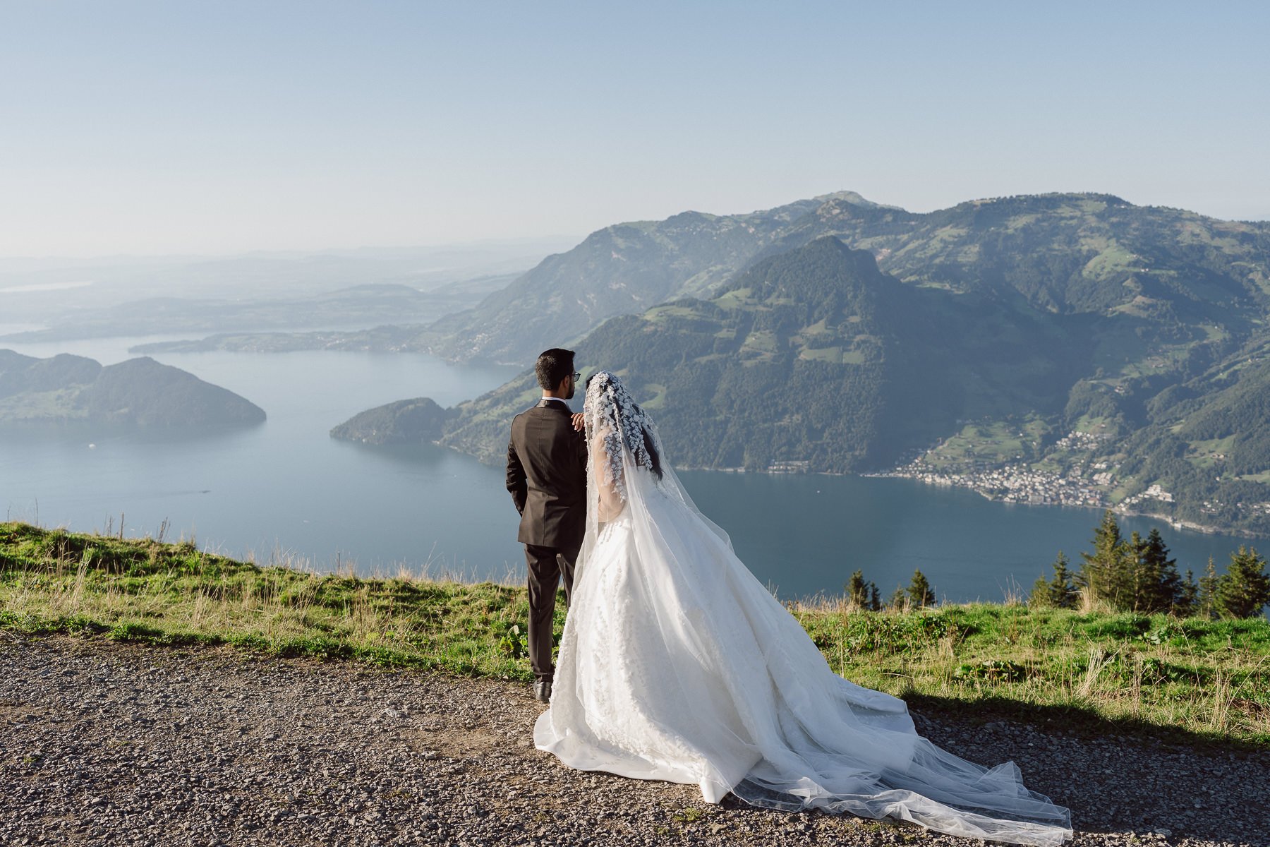 Couple de mariés face à au lac des 4 cantons lac et aux montagnes suisses, robe avec longue traîne au vent – mariage en Suisse par Nonante Émotions