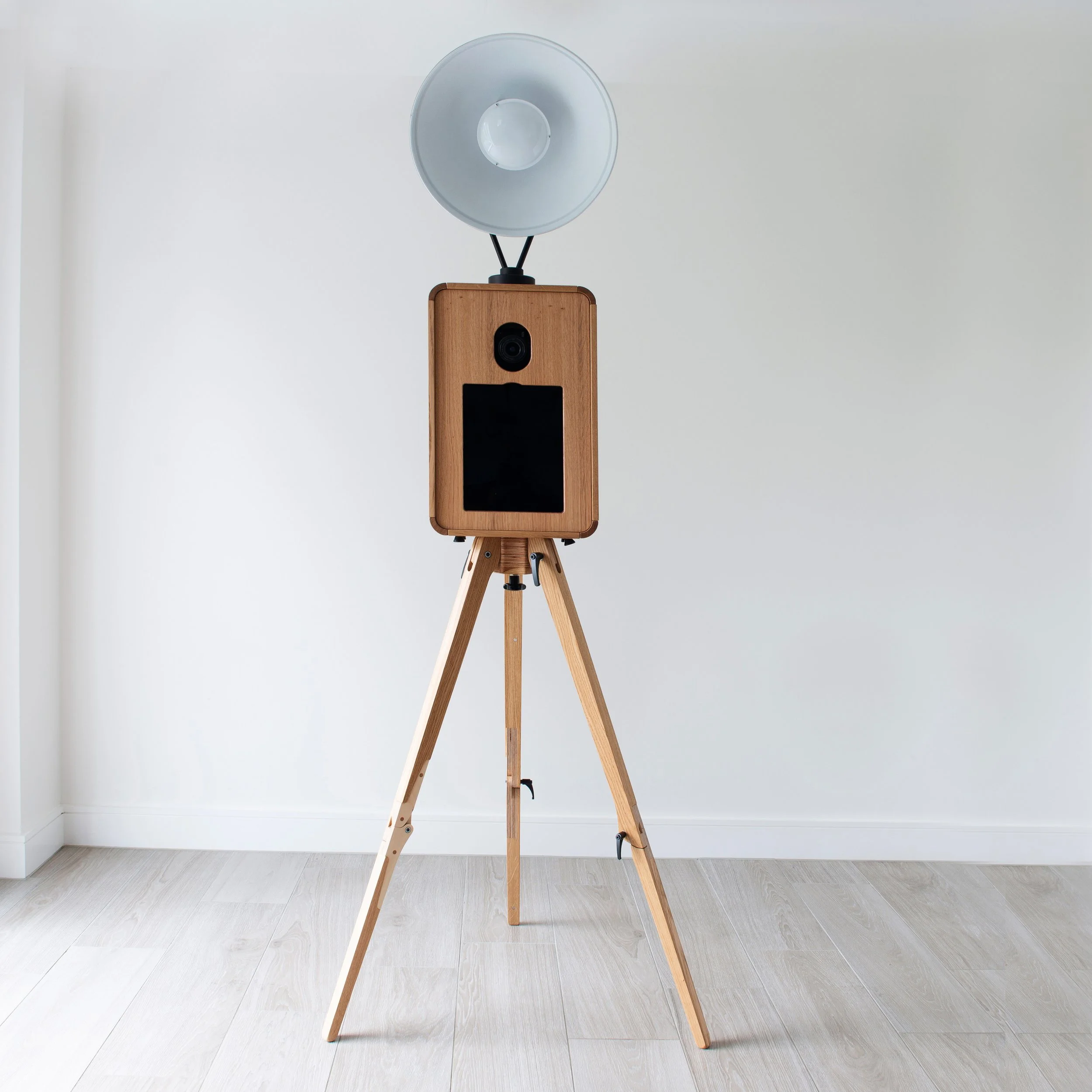 Photographie d'une caméra de sécurité en bois avec un flash à LED sur un trépied en bois dans une pièce vide avec murs blancs et plancher en bois clair.