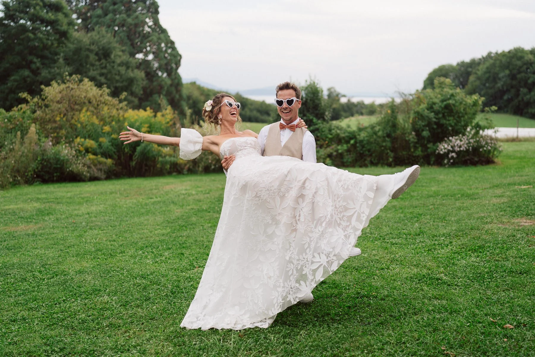 Couple de mariés riant dans un jardin verdoyant, le marié portant la mariée dans ses bras, tous deux avec des lunettes en forme de cœur, photo de mariage naturelle et joyeuse par Nonante Émotions