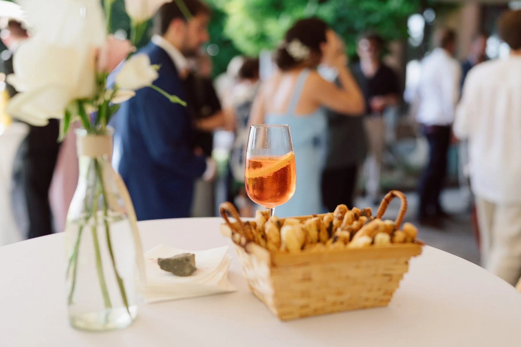 Détails du vin d’honneur de mariage avec un verre de cocktail posé sur une table, panier de biscuits et invités en arrière-plan flouté