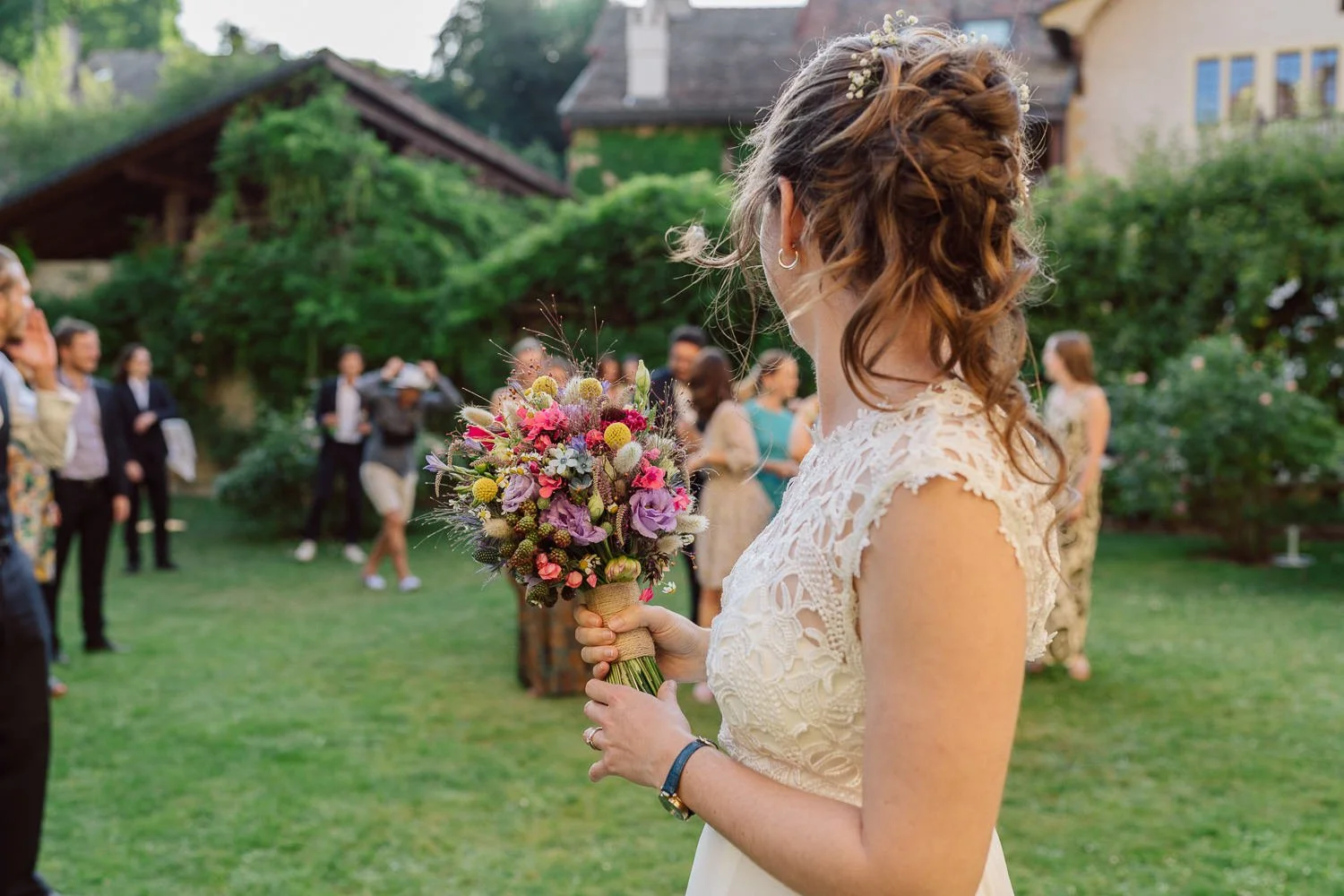 La mariée s’apprête à lancer son bouquet lors du mariage au Jardin Baillival à Romainmôtier, invités rassemblés dans le jardin, moment joyeux et spontané, photo par Nonante Émotions.