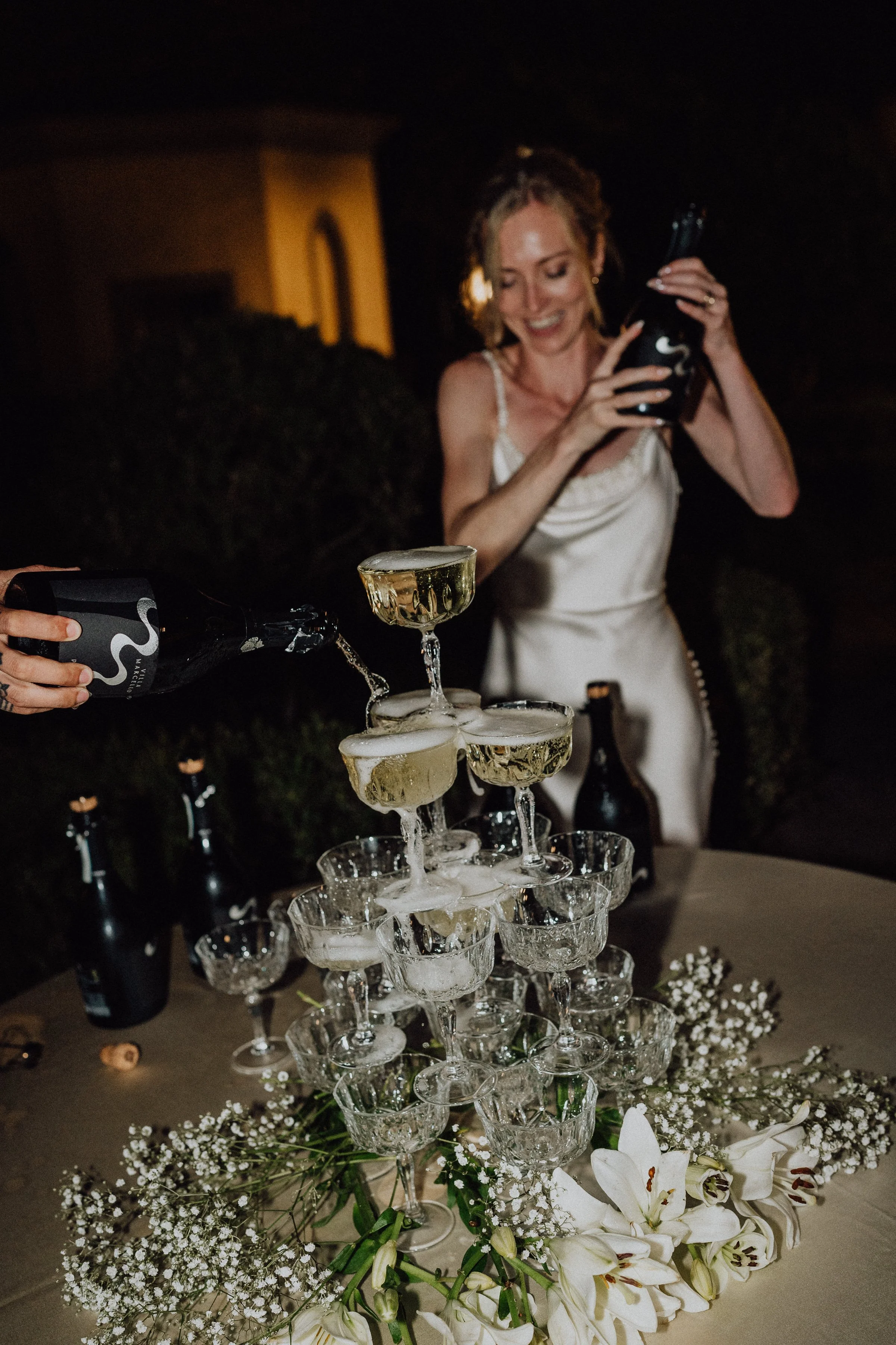 Une mariée rayonnante en robe de satin verse du champagne dans une élégante pyramide de coupes en cristal, créant une cascade festive et pétillante. La table est décorée de gypsophile et de lys blancs, ajoutant une touche de douceur à ce moment de cé