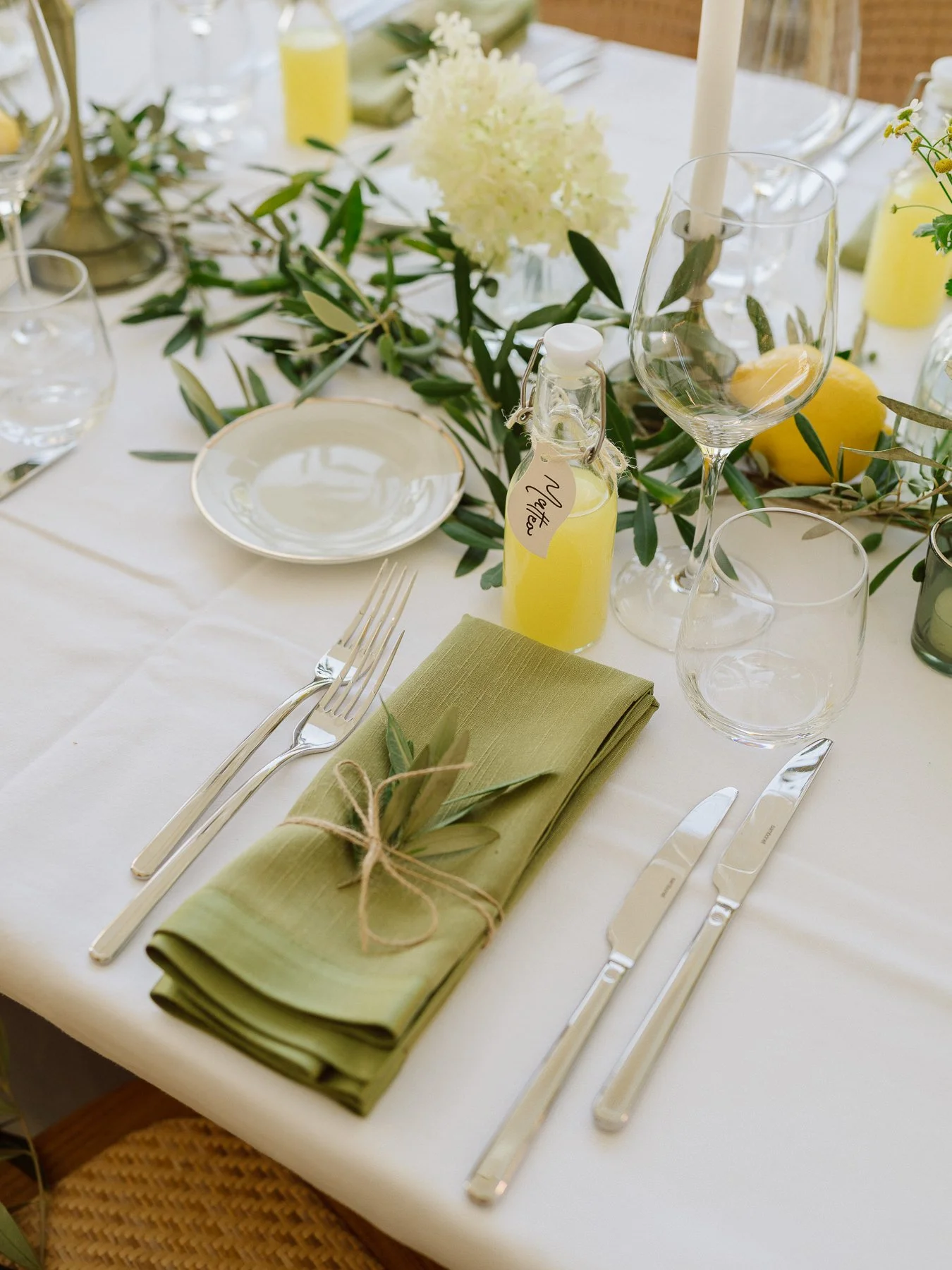 Détail d’une table de mariage en Toscane avec serviette vert sauge, branche d’olivier, citrons, fleurs blanches et jaunes et petits flacons d’huile d’olive en cadeau invité.