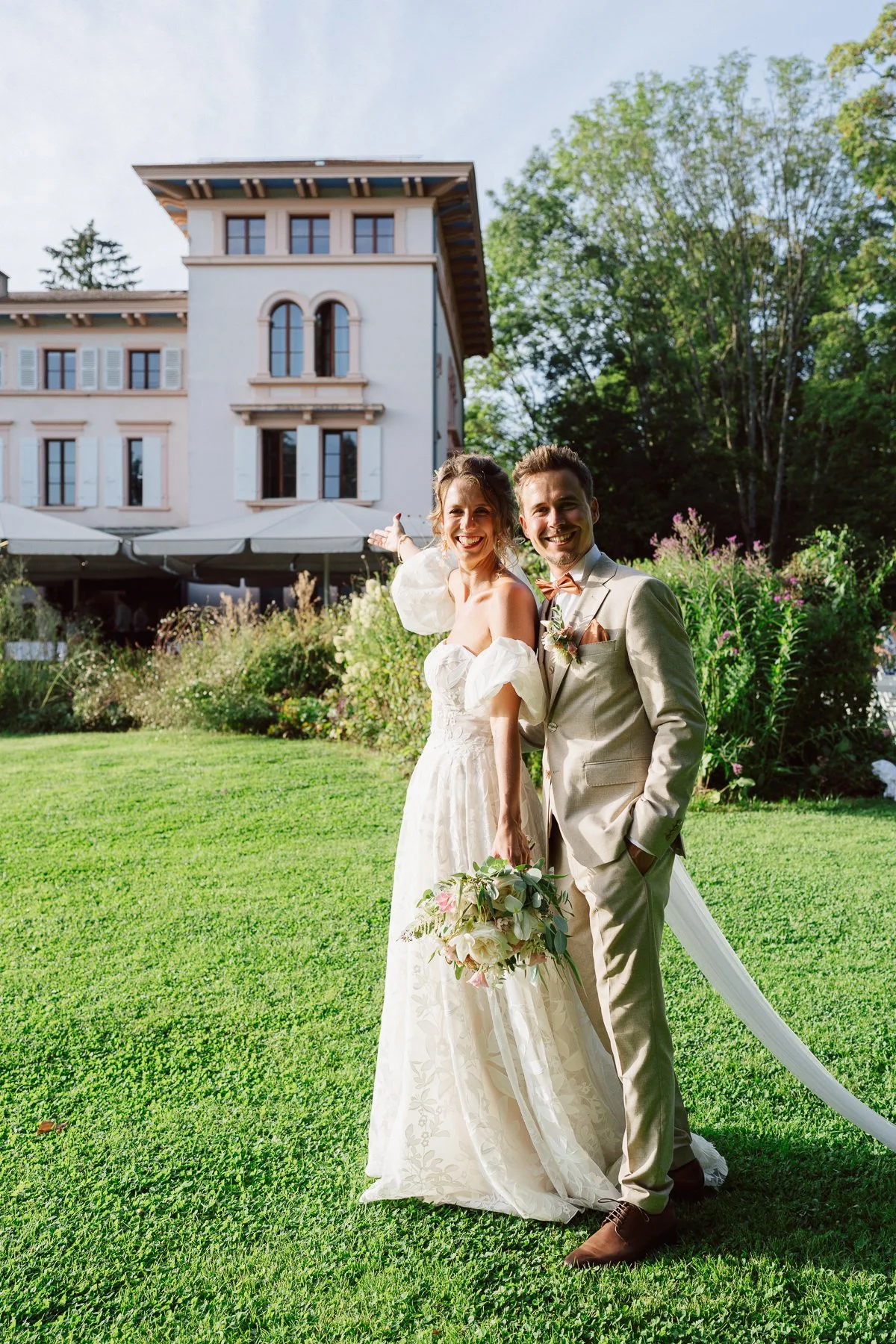 Couple de mariés souriants dans le jardin d’un château, la mariée présentant le lieu de réception derrière eux, photo de mariage lumineuse et naturelle réalisée par Nonante Émotions
