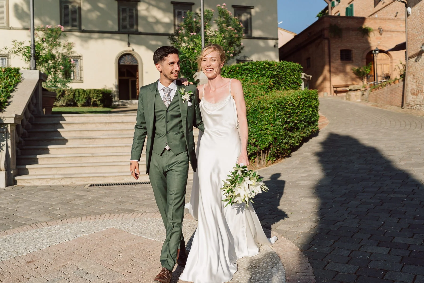 Jeunes mariés marchant bras dessus bras dessous dans un village toscan ensoleillé, la mariée tenant un bouquet de fleurs blanches et le marié portant un costume vert élégant.