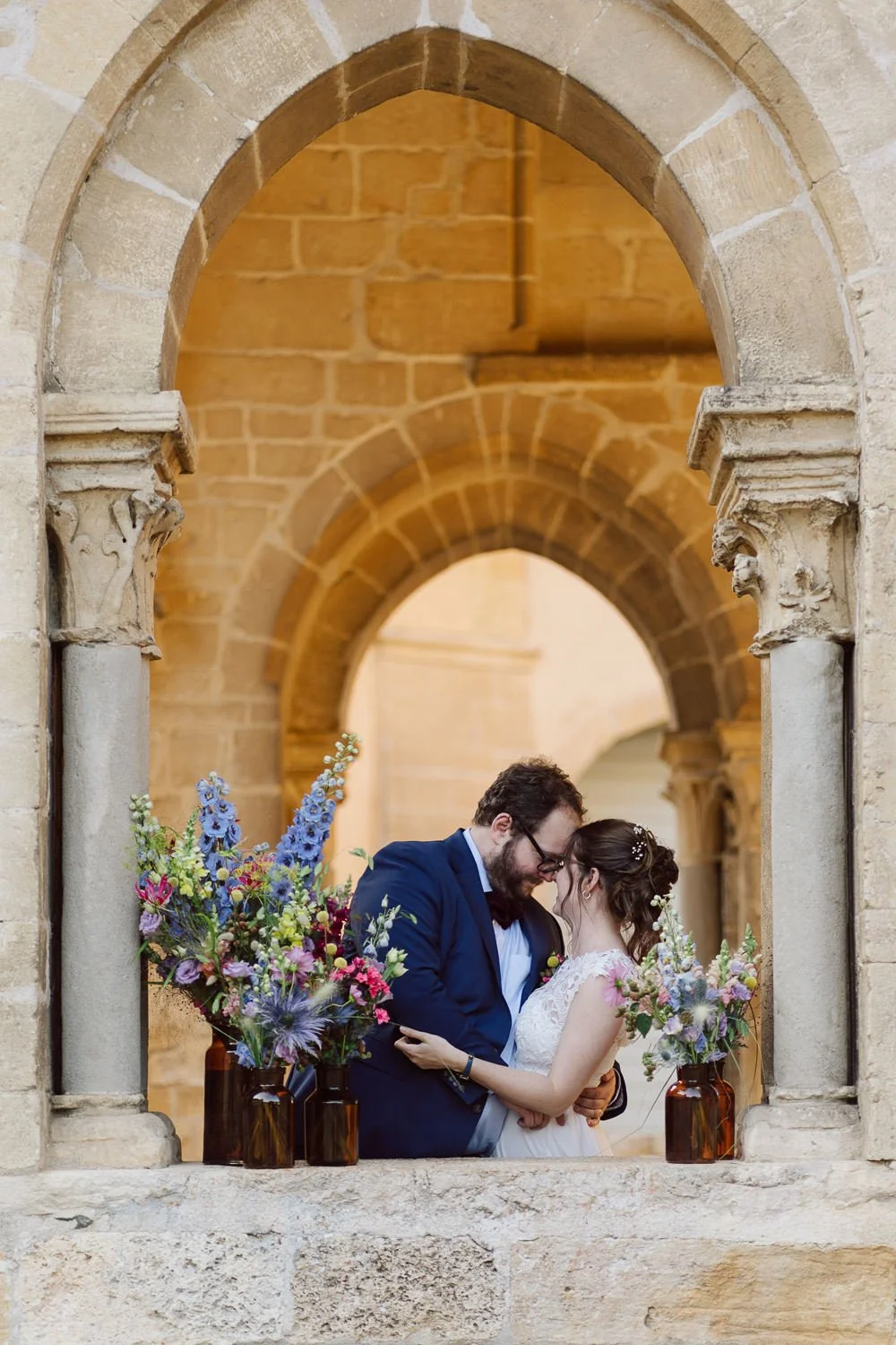 Un couple marié partage un moment intime à travers une fenêtre en pierre antique, entouré de bouquets de fleurs colorées dans des vases en verre.