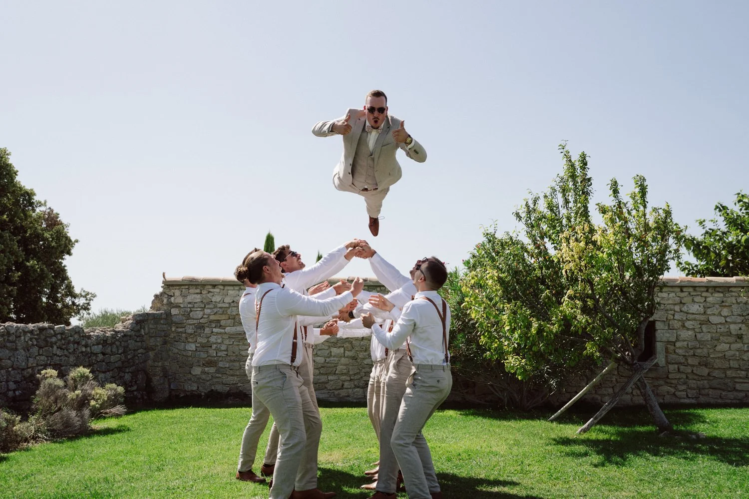 Le marié en costume beige lancé en l’air par ses garçons d’honneur dans un jardin en pierre sous le soleil, scène fun et dynamique de mariage en Provence.