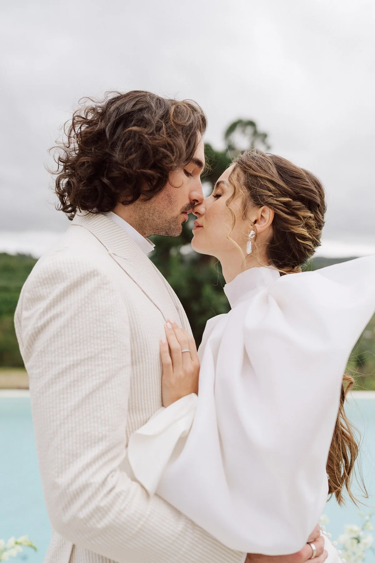 Un baiser passionné entre les mariés dans un cadre romantique près d’une piscine. La mariée, habillée d’une robe minimaliste et élégante, et le marié, vêtu d’un costume clair, incarnent le raffinement et l’intimité.