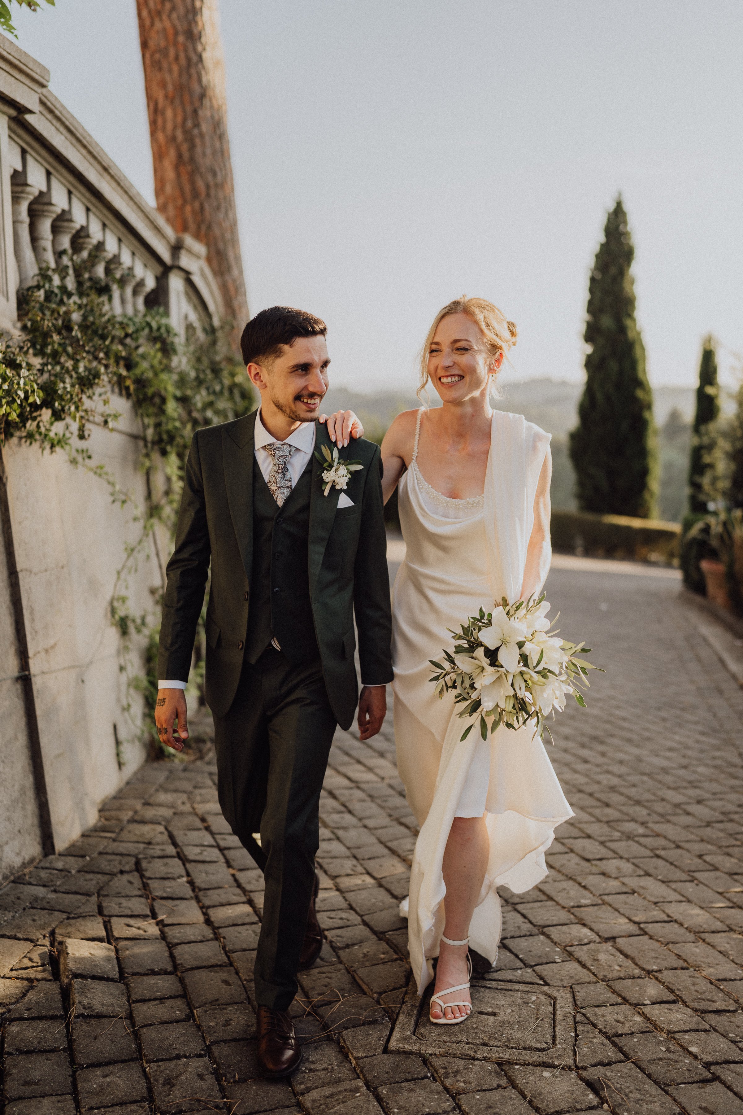 Un couple de mariés rayonnant marche ensemble dans le cadre romantique de la Toscane, au domaine de Borgo Bucciano, entouré de verdure et d’une architecture élégante en pierre. La mariée porte une robe fluide accompagnée d’un bouquet de fleurs blanch