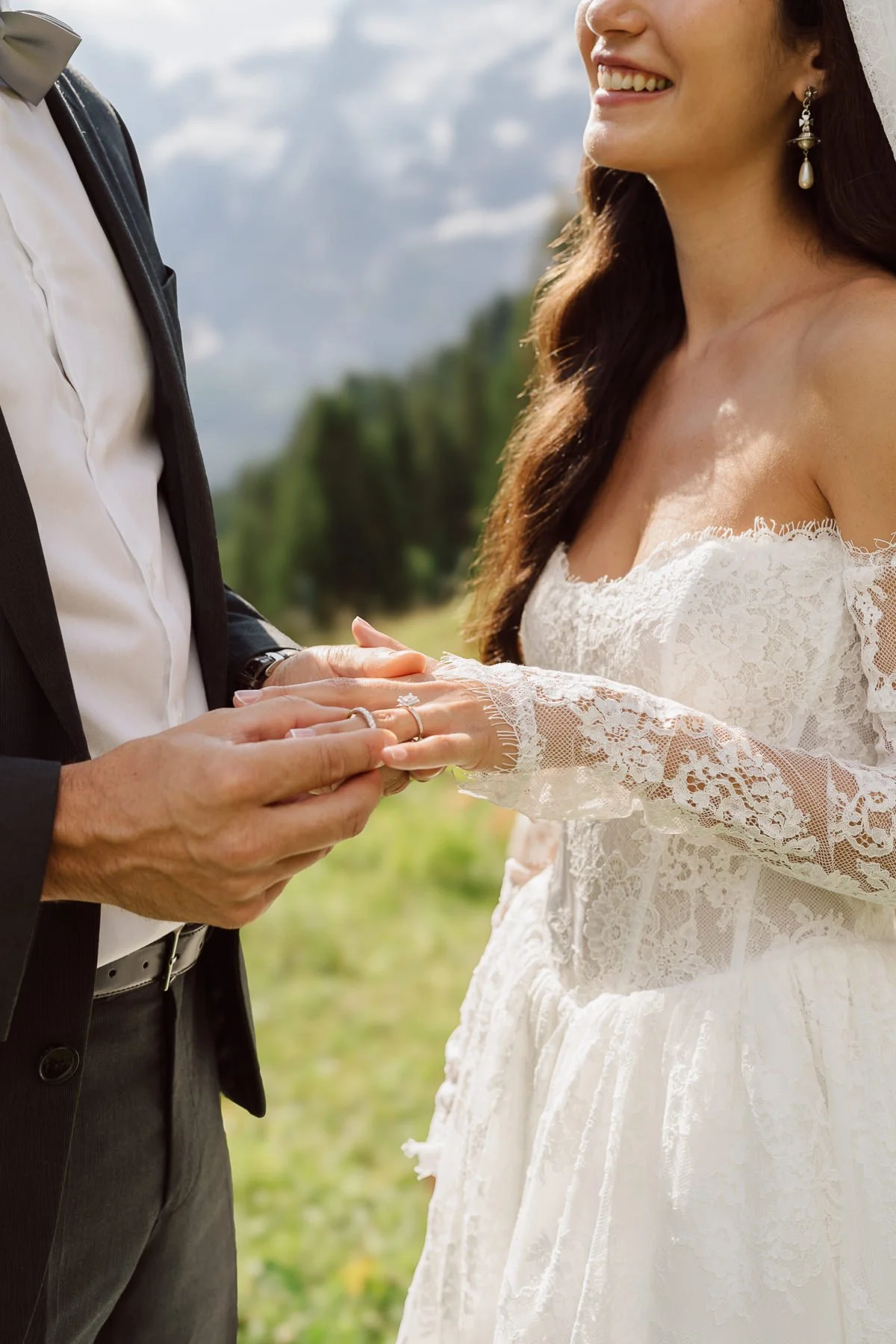 Un couple marié échange des alliances en plein air, la femme porte une robe en dentelle blanche et des boucles d'oreilles pendantes, l'homme porte un costume noir et une chemise blanche. La scène se déroule dans un paysage naturel verdoyant.