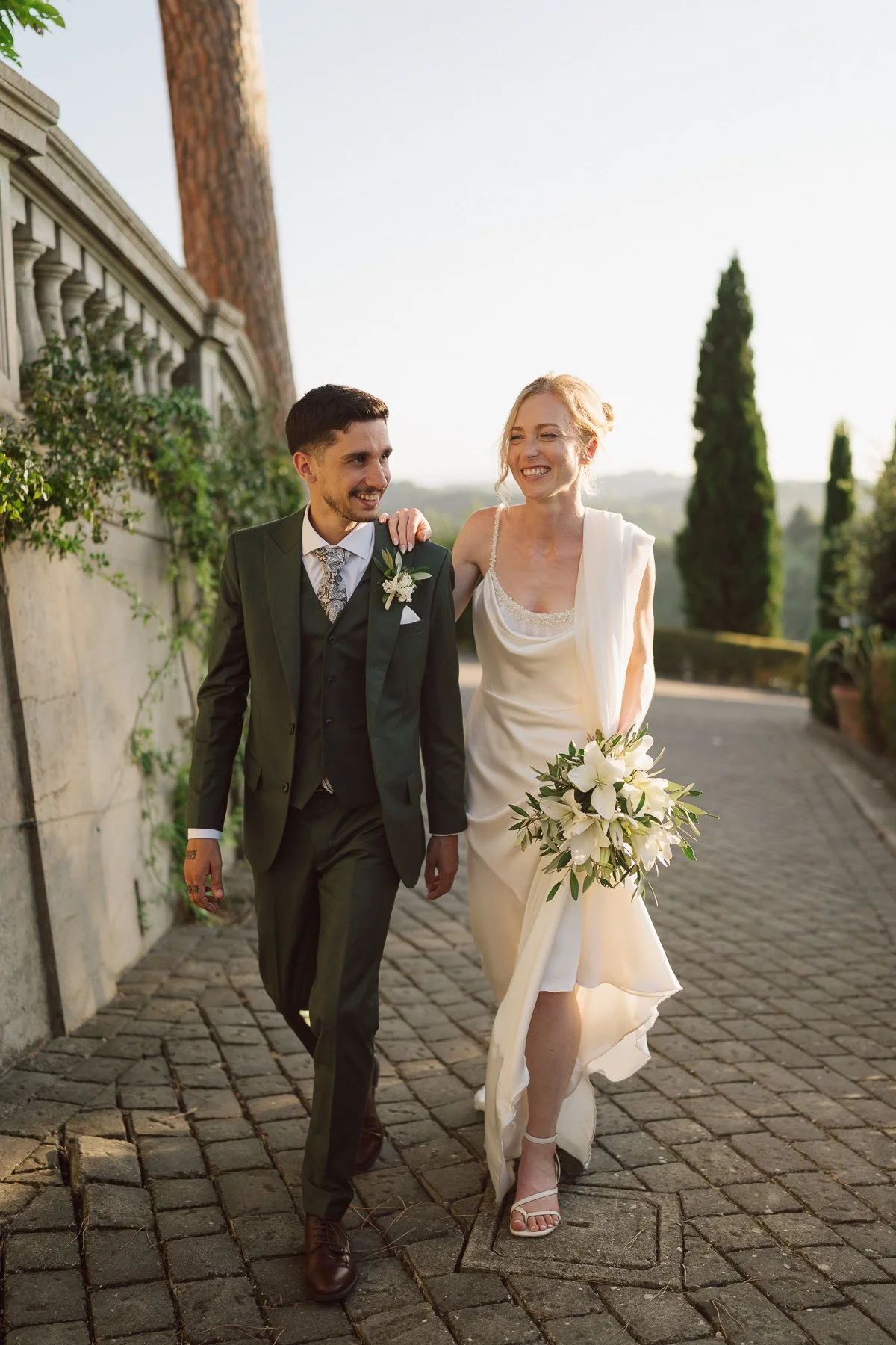 Jeunes mariés marchant côte à côte au coucher du soleil en Toscane, la mariée en robe fluide tenant un bouquet blanc, le marié en costume vert, ambiance douce et romantique.