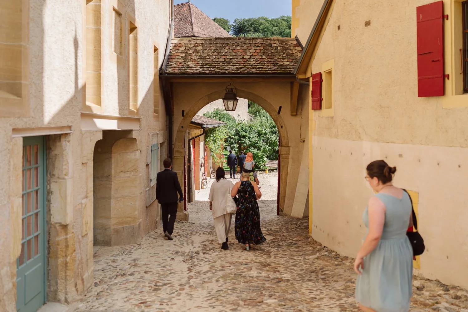 Invités de mariage marchant sous les arcades du Jardin Baillival à Romainmôtier, ruelle pavée et architecture historique, ambiance chaleureuse et authentique, photo par Nonante Émotions.