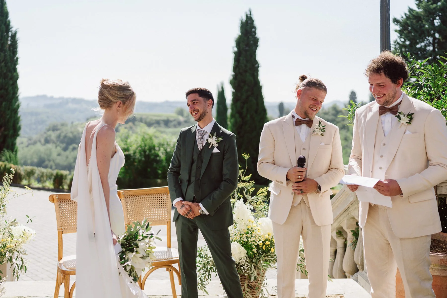 Cérémonie de mariage en plein air avec la mariée en robe blanche et trois hommes en costumes clairs ou foncés, souriants, lors d'une journée ensoleillée avec un décor verdoyant en arrière-plan.