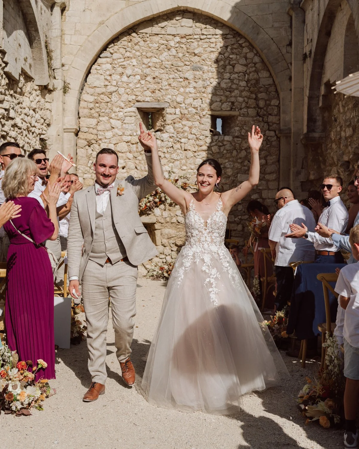 Les mariés sortent de leur cérémonie laïque sous une arche en pierre, bras levés de joie, pendant que leurs invités les applaudissent dans une cour en pierre en Provence au domaine de Sarson