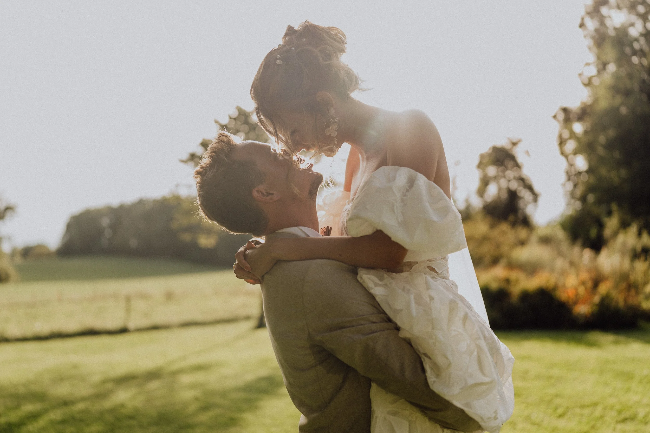  Un moment joyeux où le marié soulève sa mariée sous la lumière douce de la fin de journée. La robe volumineuse de la mariée et leur éclatante complicité captivent dans un cadre bucolique ensoleillé.