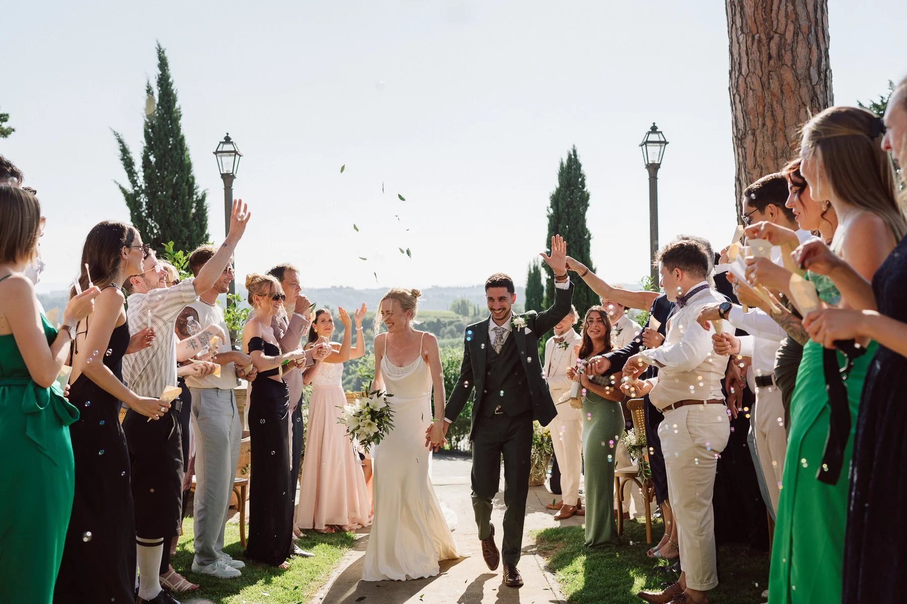 Sortie de cérémonie de mariage en Toscane, mariés marchant sous une pluie de confettis et d’olivier lancés par les invités en extérieur