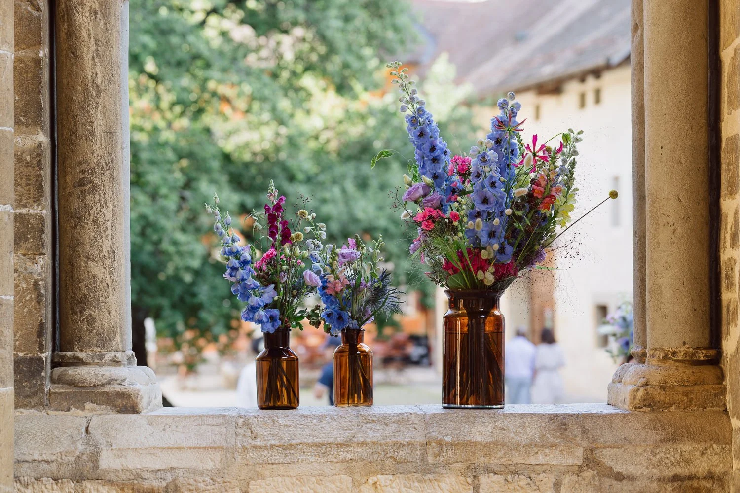 Compositions florales colorées dans des vases en verre ambré posées sous les arcades du Jardin Baillival à Romainmôtier, décoration de mariage élégante et naturelle, photo par Nonante Émotions.