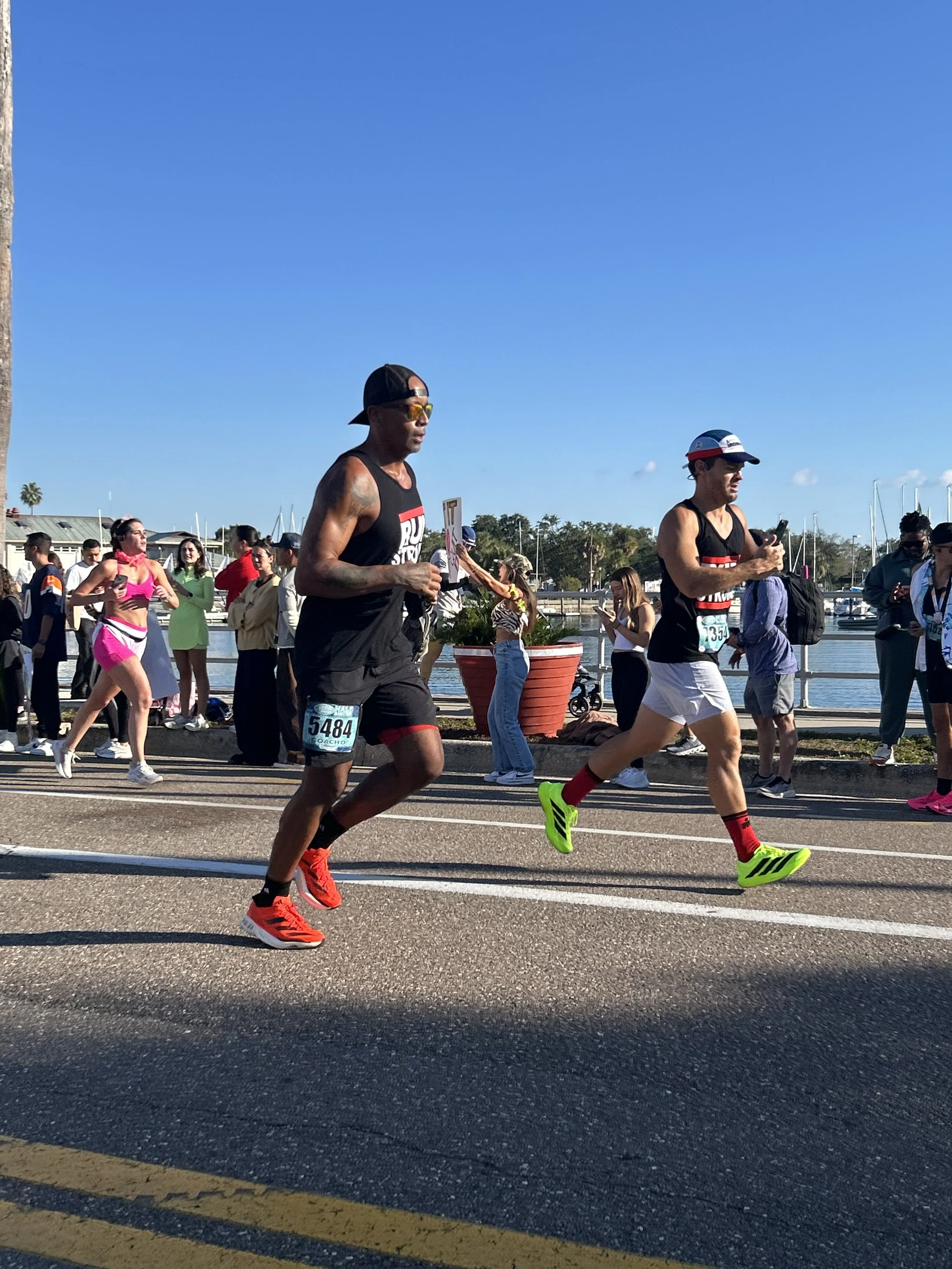 Two runners participating in the St Pete RUN Fest half marathon, running on a road near St Petersburg marina with boats, while spectators watch from the sidewalk.