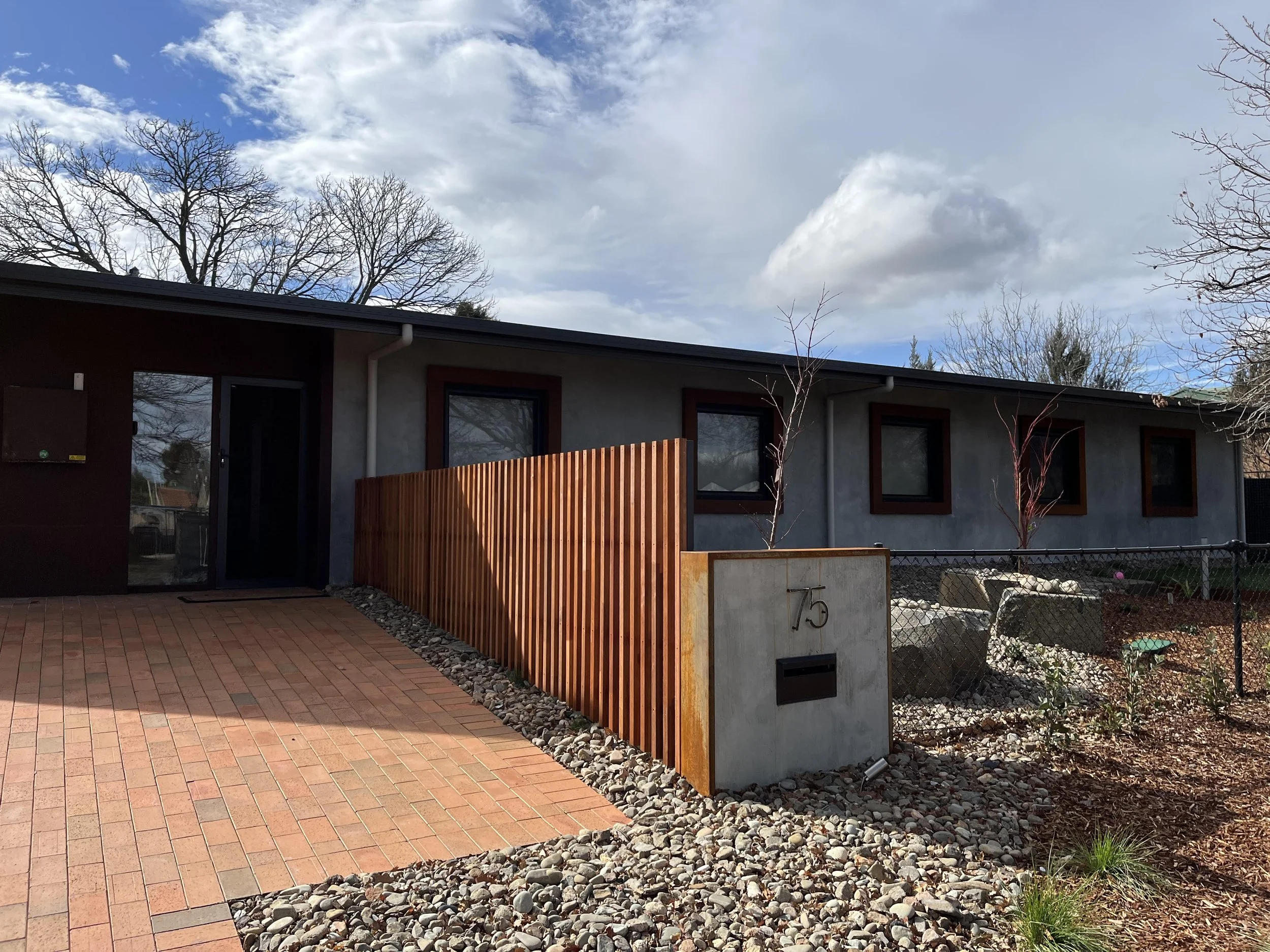 Photo of a modern house with gray and red exterior walls, three windows, and a wooden fence. The yard has a paved brick patio and a gravel border. The sky is partly cloudy with some trees in the background.
