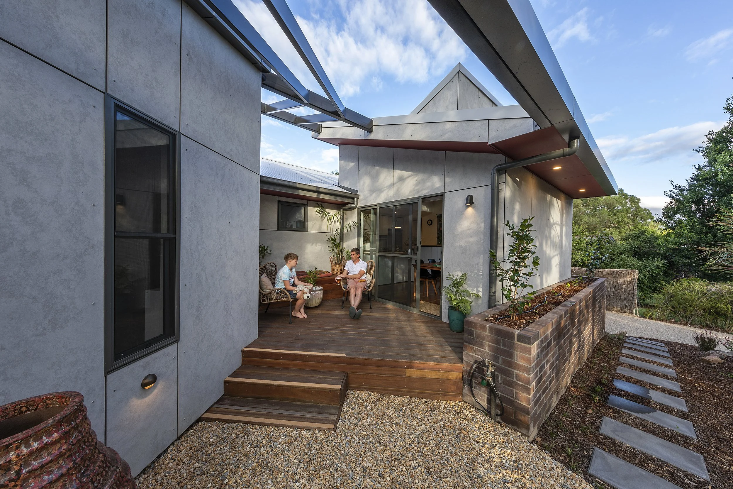 Modern home with a patio where two people are sitting and talking, surrounded by plants and greenery, with a blue sky and clouds overhead.