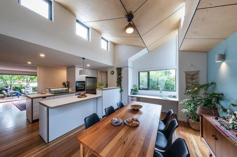 Modern dining area with a wooden table, black chairs, and large windows showing outdoor greenery. Open kitchen with white cabinets, a countertop island, and plants, with additional large windows and a sitting area visible in the background.