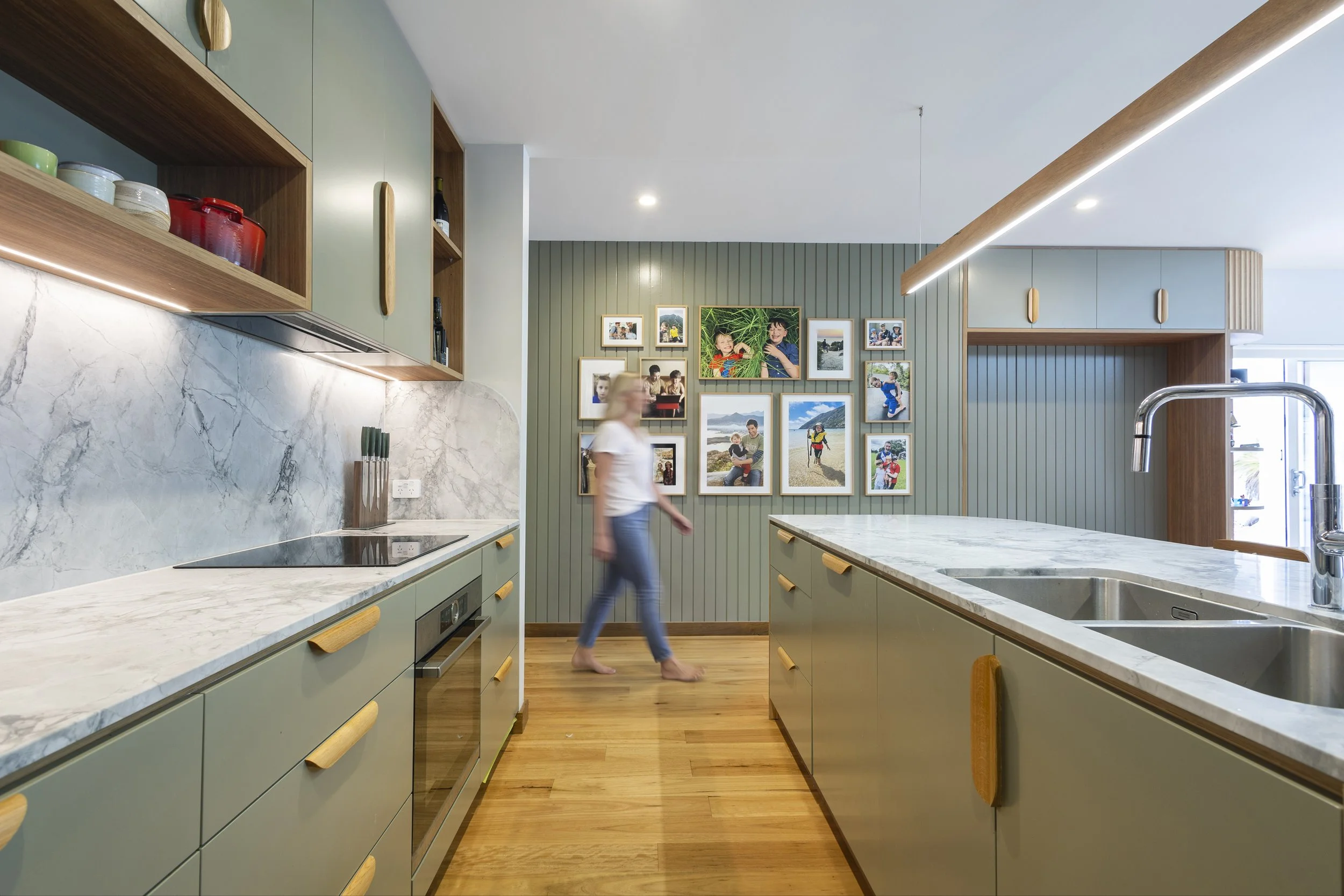 Modern kitchen with green cabinetry, marble countertops, and a gallery wall of family photos. A woman walking barefoot past the kitchen island.