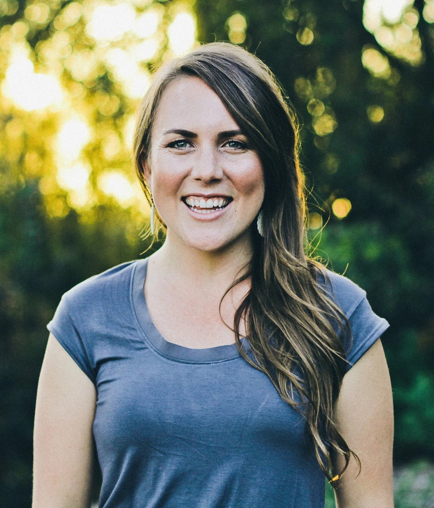 A young woman with long wavy brown hair and light skin smiling outdoors during sunset, wearing a blue t-shirt, with blurred yellow-green foliage and sunlight in the background.