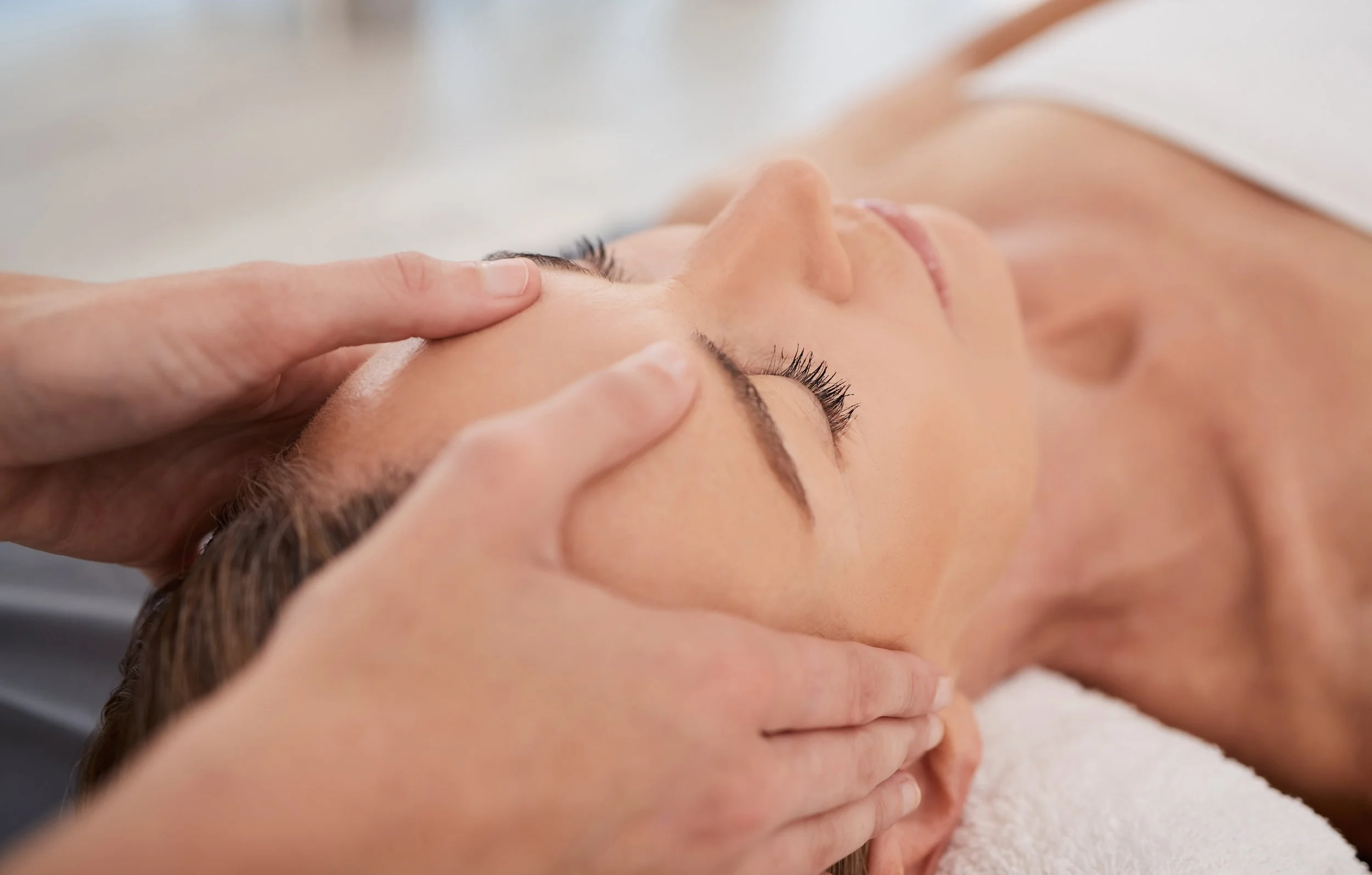A woman lying face up on a massage table receiving a massage, with care provider's hands on her forehead and face.