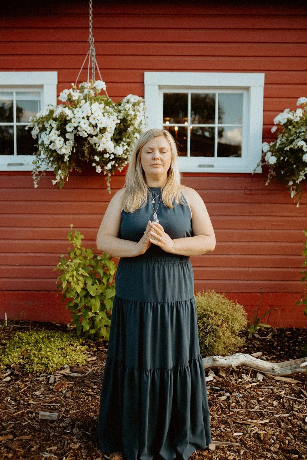 Person in a navy dress meditating outdoors in front of a red building with white hanging flowers and windows.