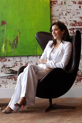 Queen Mary of Denmark  wearing a white suit sitting in a black modern lounge chair against a textured white brick wall with a large green abstract painting.