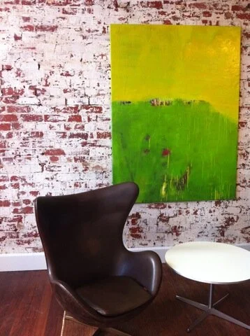 A modern black lounge chair with a rounded design, next to a small white side table with a metal base, in front of a brick wall with a large abstract painting in yellow and green.