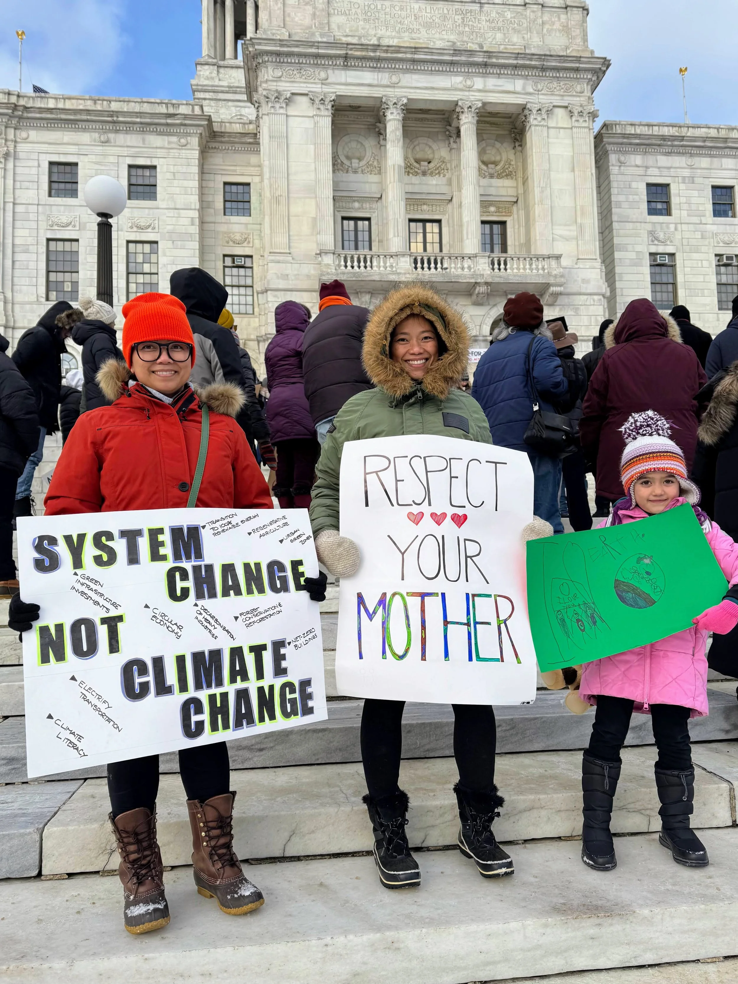 Three people, two women and a girl, standing on city steps holding signs at a climate change protest. The woman on the left holds a sign that reads "System Change Not Climate Change," the woman in the middle holds a sign that says "Respect Your Mother," and the girl on the right holds a green poster. They are dressed warmly in winter clothing, and behind them is a large white government building with many people gathered around.