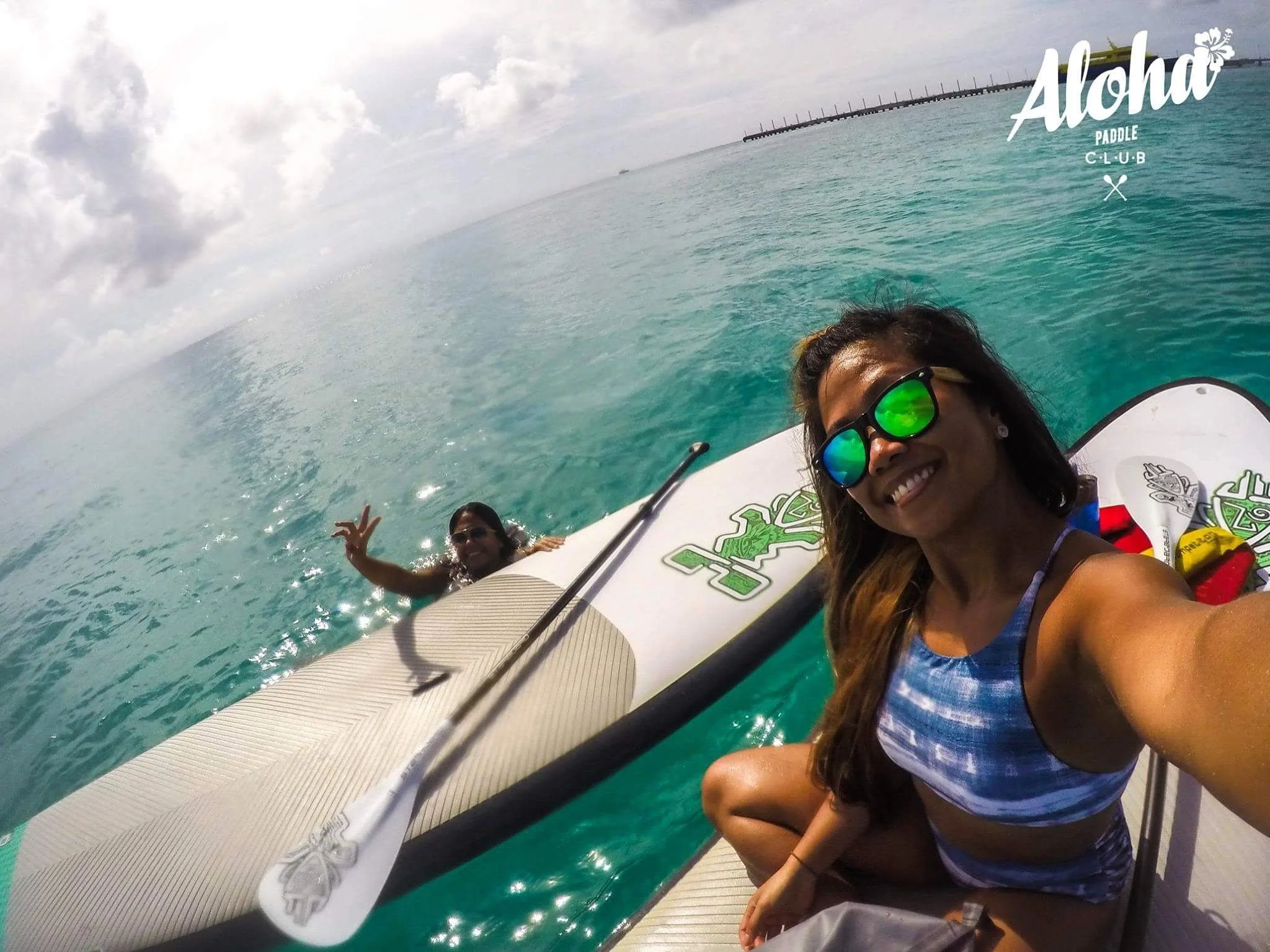 Smiling woman with sunglasses taking a selfie on a paddleboard in the ocean, with two other women in the background, one making a peace sign, under cloudy sky near a pier.