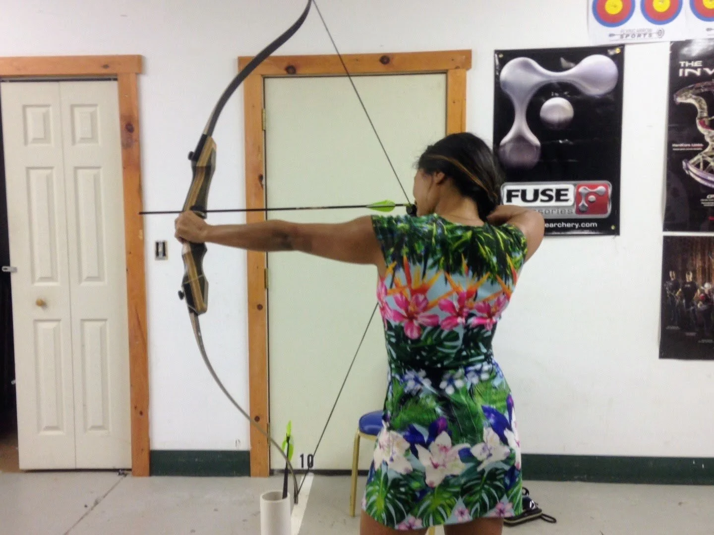 Woman practicing archery indoors, aiming a bow and arrow with her right arm extended.