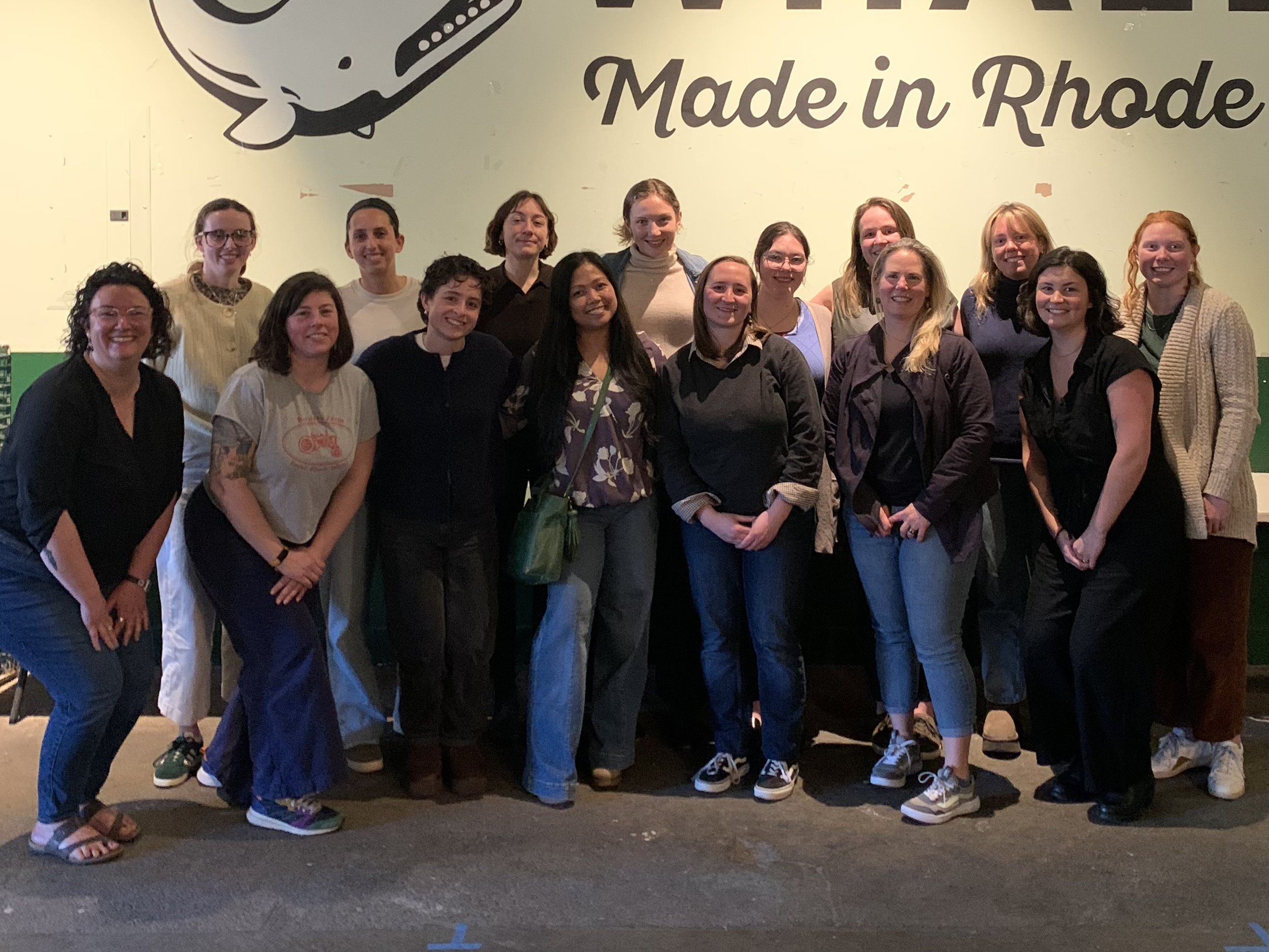 Group of fifteen women standing in front of a wall with the words 'Made in Rhode' visible. The women are smiling and dressed casually.