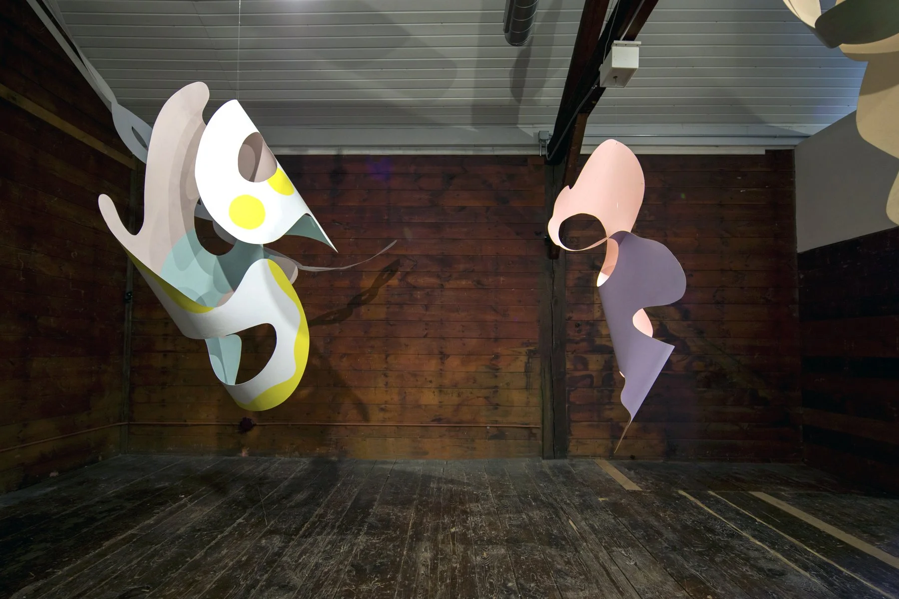 Two abstract, colorful hanging sculptures with geometric shapes and smooth curves inside a room with wooden walls and a wood floor.