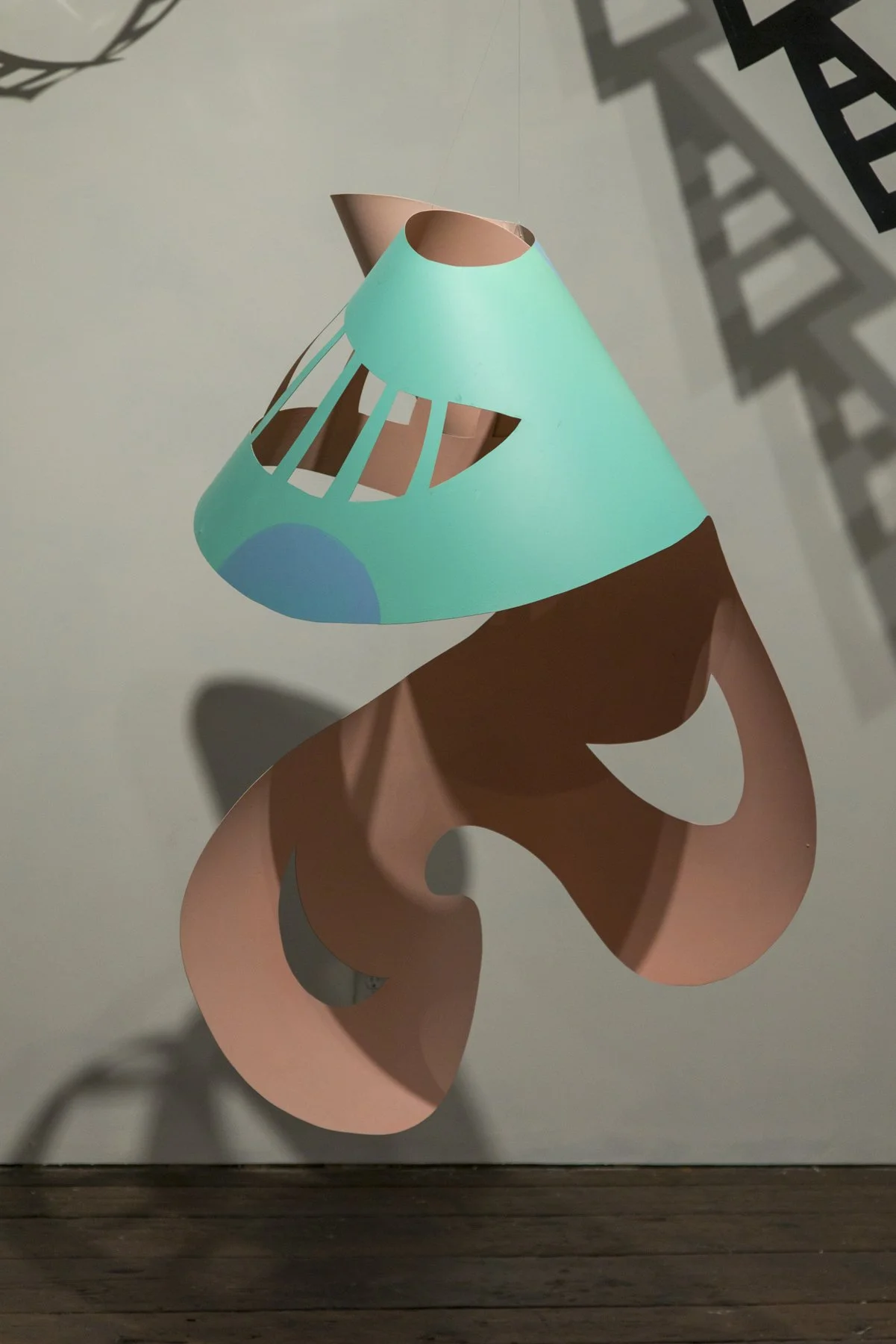 An abstract sculpture resembling a lamp, with a turquoise blue lampshade and a pink base, hanging in a gallery space.