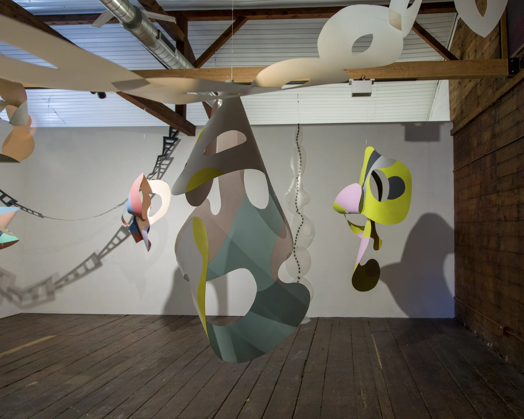 Colorful abstract paper sculptures hanging from the ceiling in an art installation, with shadows cast on the wooden floor and white walls.