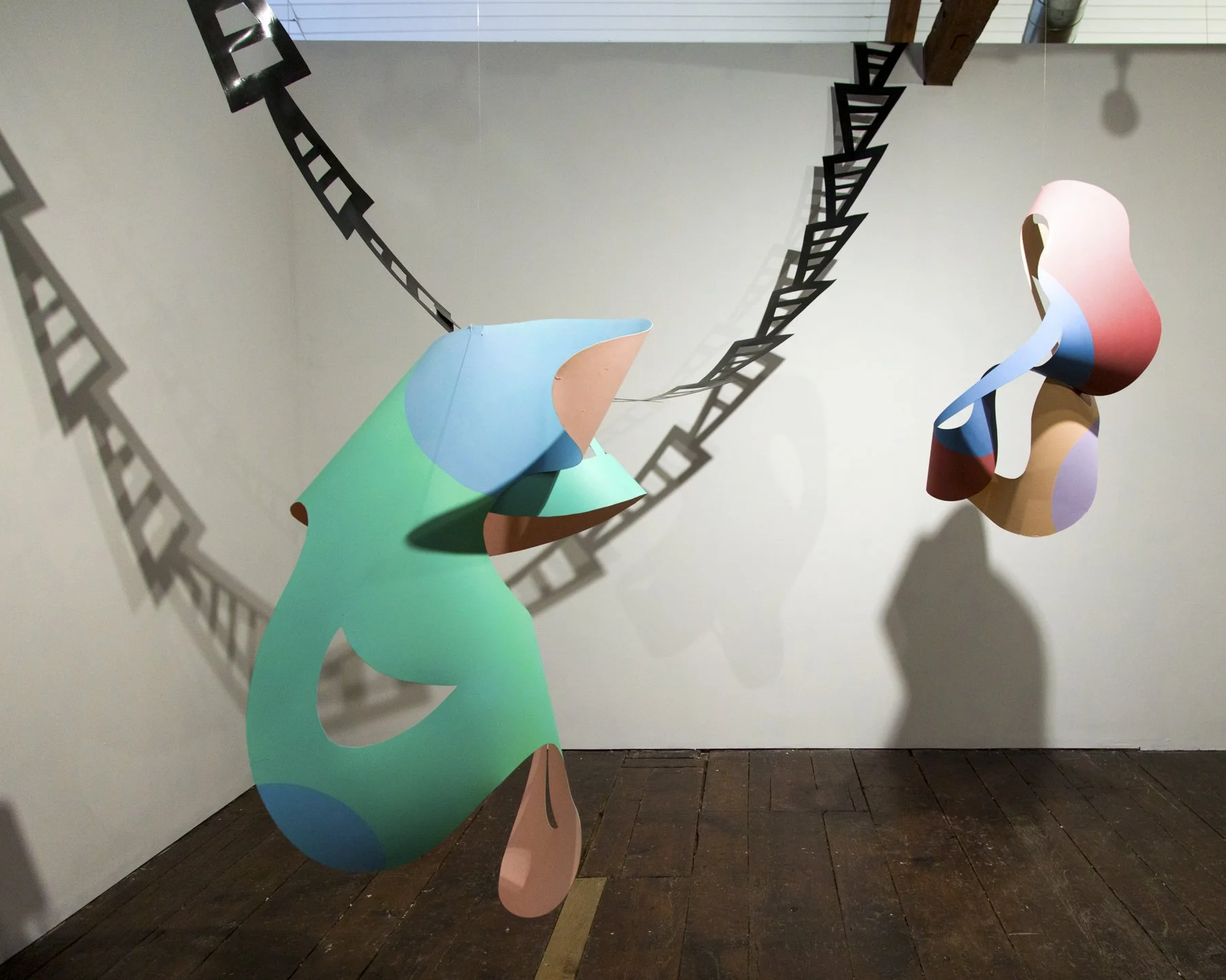 Colorful abstract sculptures hanging in an art gallery with shadows cast on the white wall behind them.