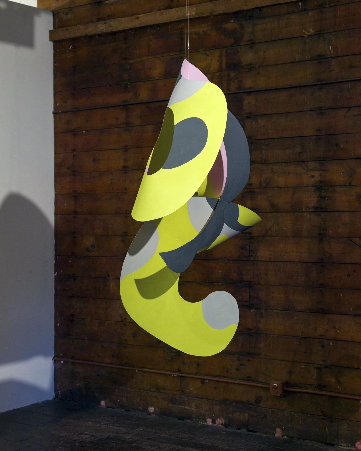 Abstract hanging sculpture with yellow, black, white, pink, and green shapes against a wooden wall.