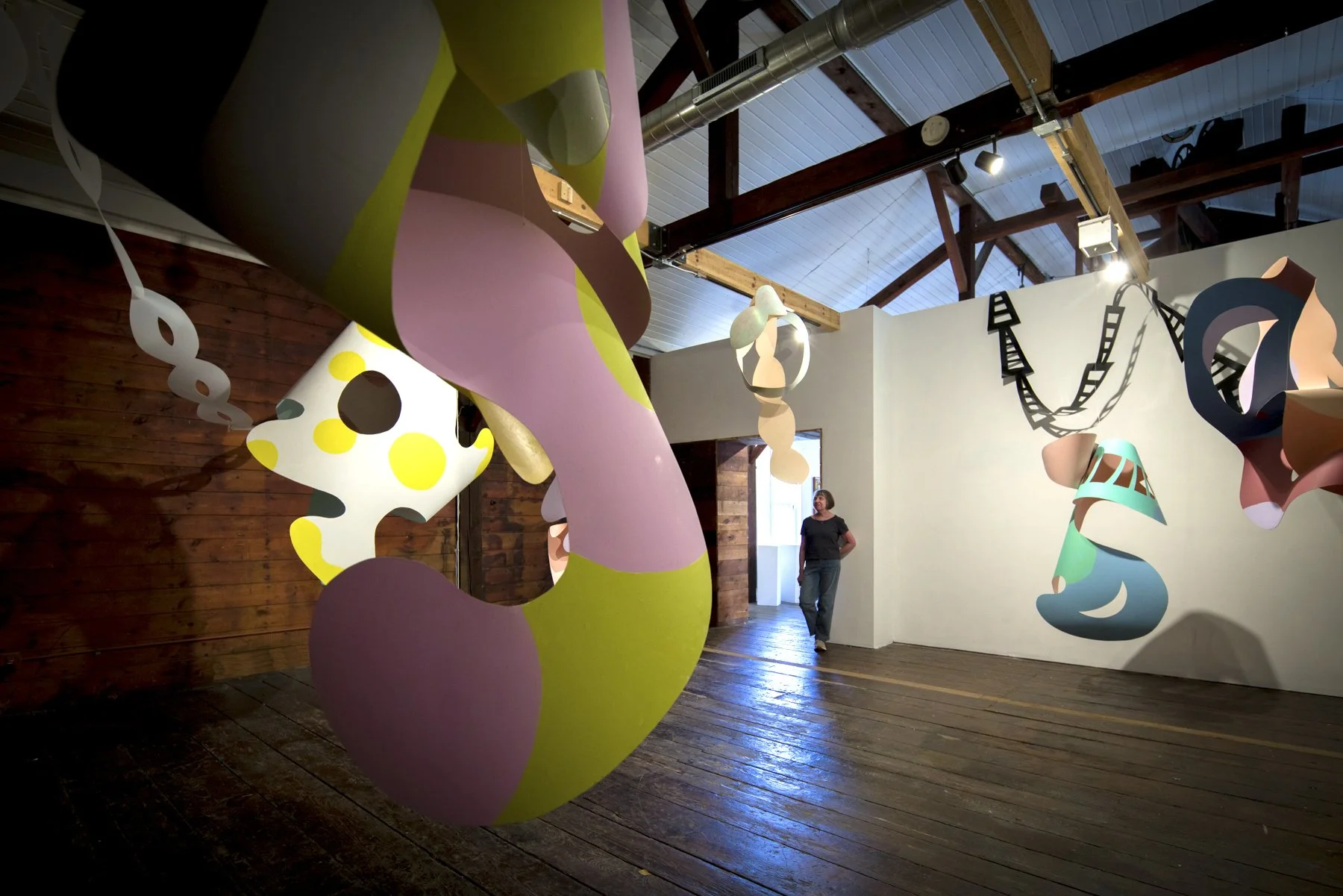 Contemporary art installation with colorful, abstract sculptures hanging from the ceiling in a gallery space with wooden floors and walls.