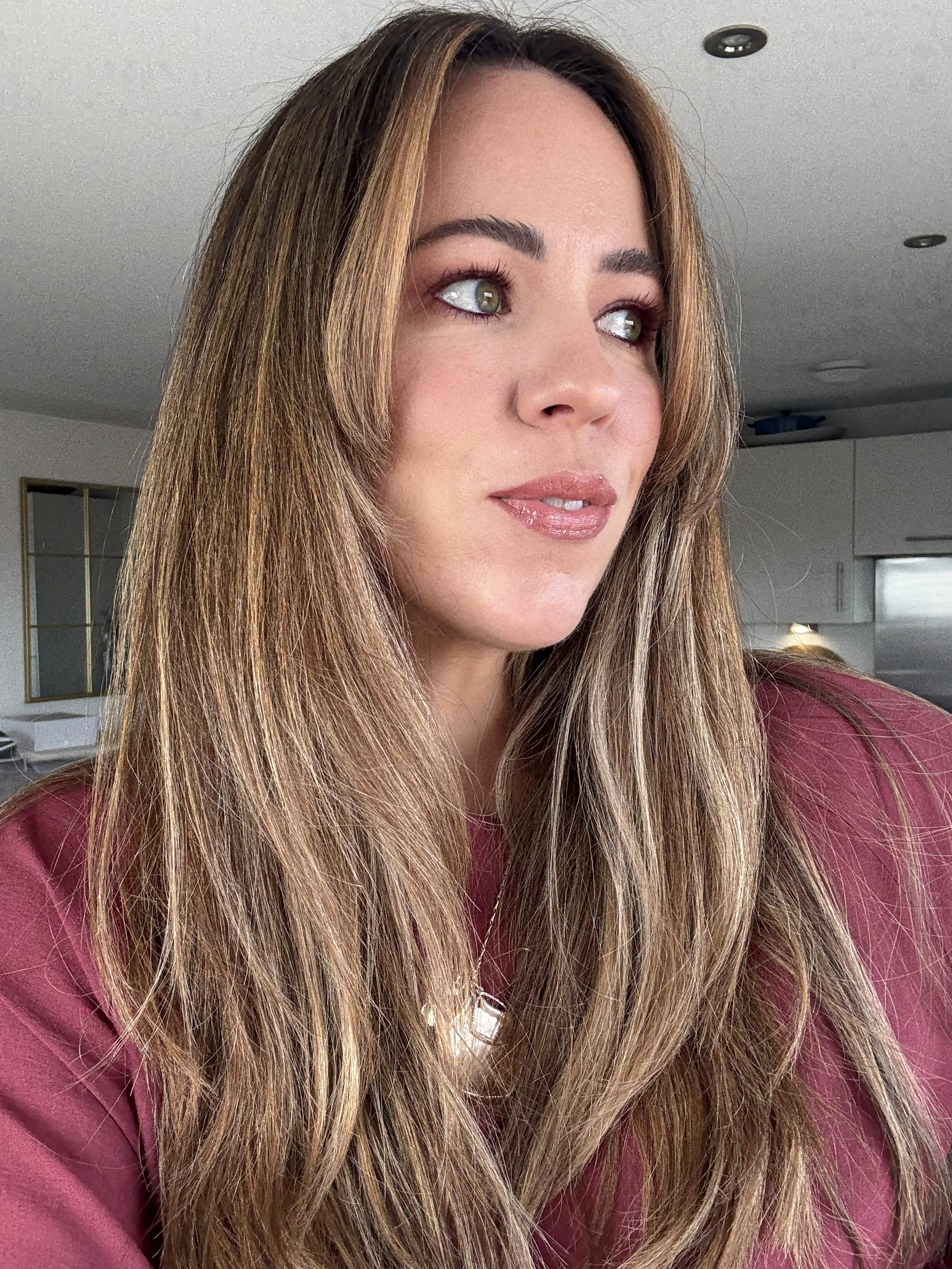 Close-up of a woman with long, wavy brown hair, wearing a maroon top, looking off to the side with a slight smile. Indoor background with white cabinets and ceiling lights.
