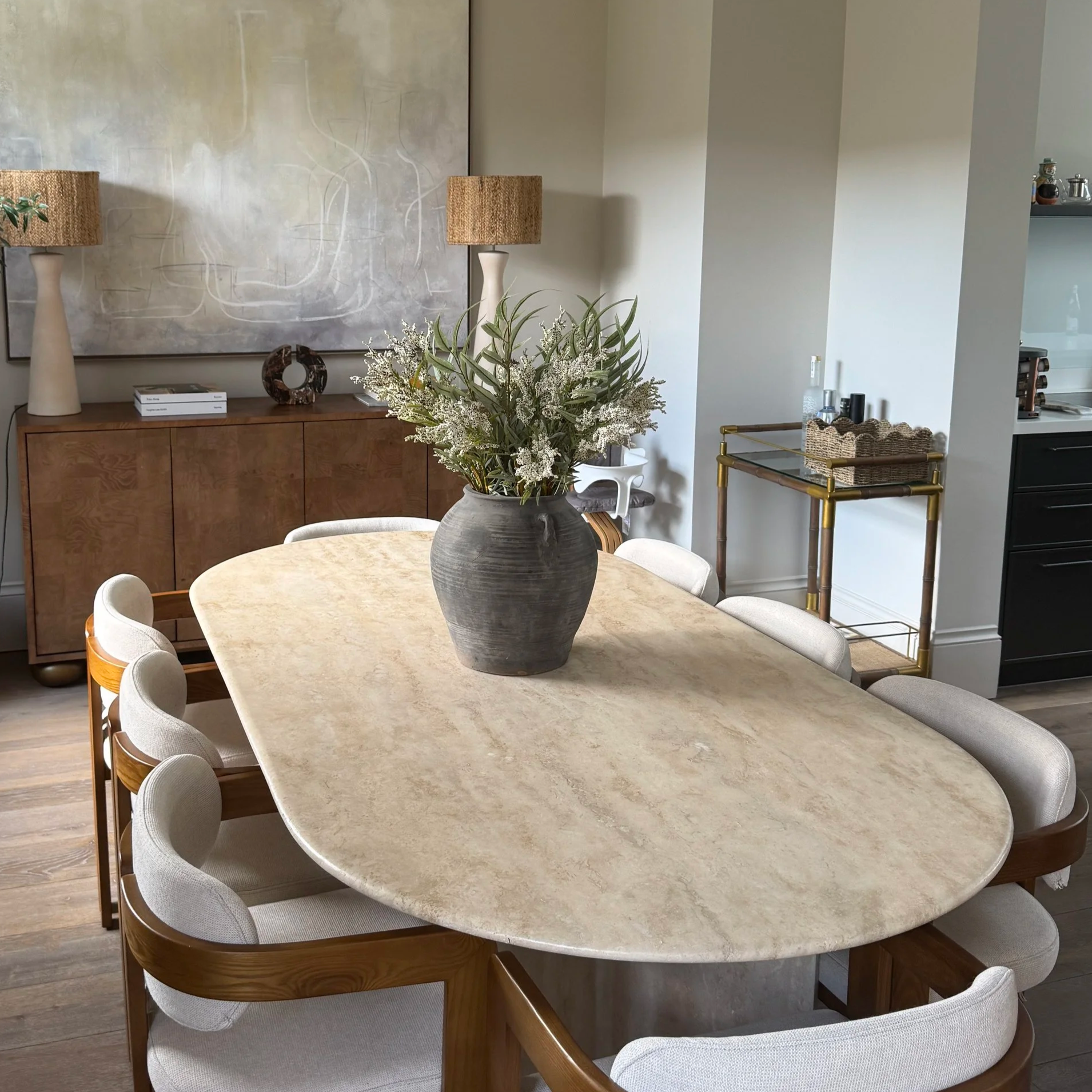 A marble dining table surrounded by8 boucle white and wood chairs, olive plant in pot with wooden sideboard and two lamps