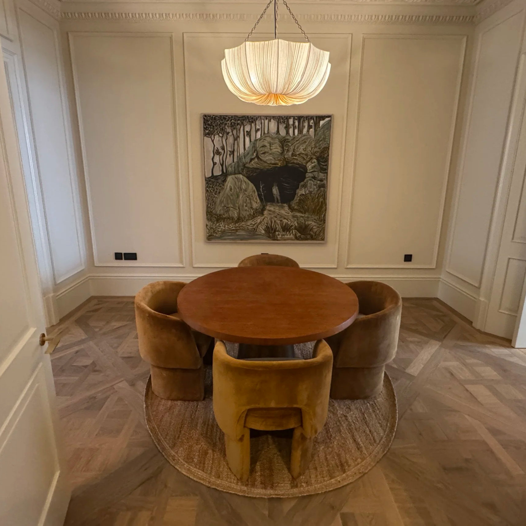 A dining room with a round wooden table surrounded by five tan velvet chairs, a textured round rug underneath, and a large painting of a cave entrance with trees and rocks on the wall behind.
