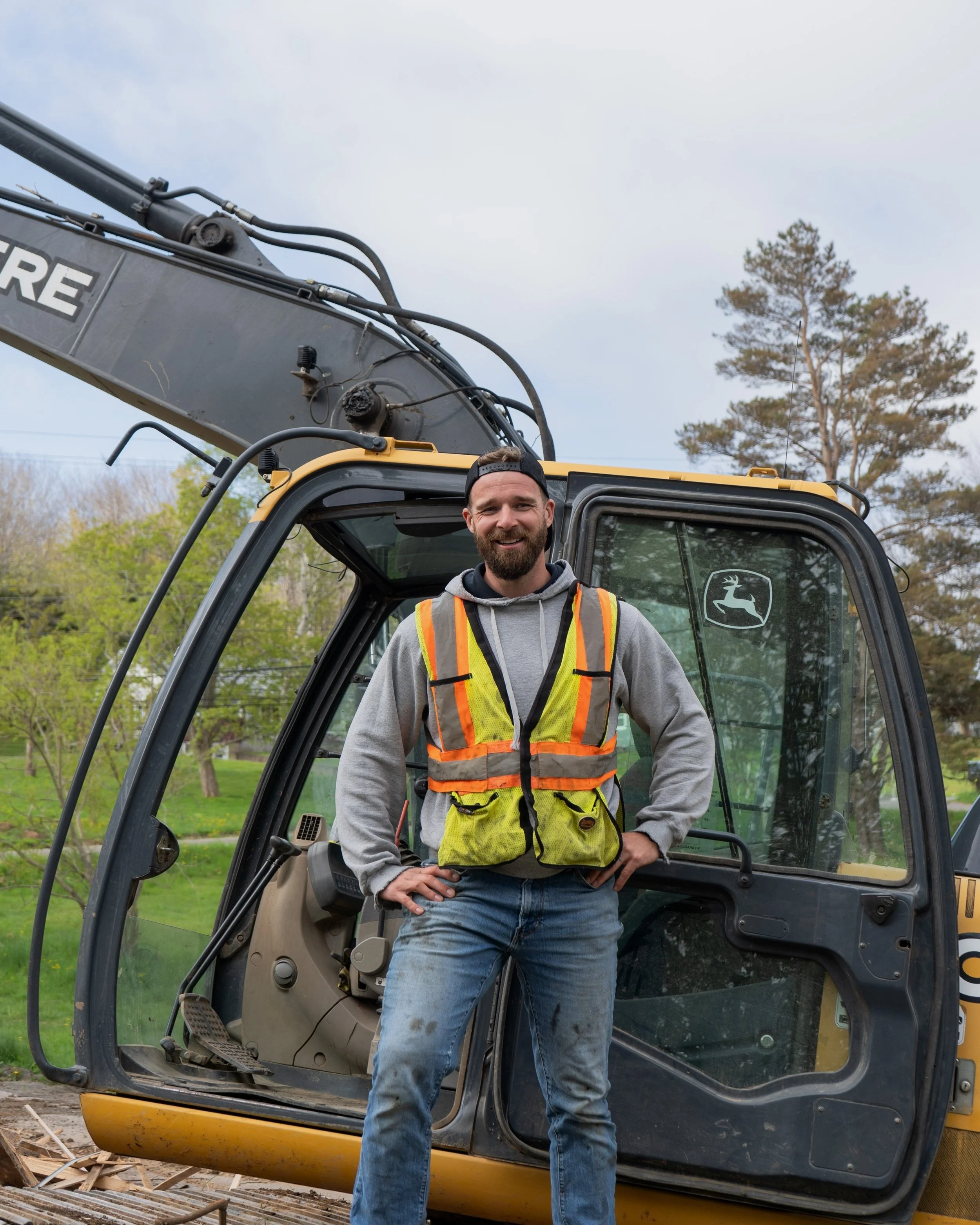 A man in a gray hoodie and high-visibility safety vest standing in front of a John Deere excavator outdoors with trees and cloudy sky background.